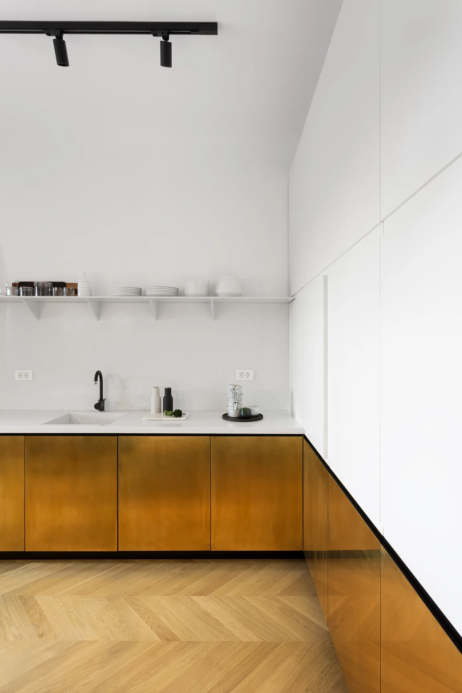 A minimalist kitchen features brass lower cabinets, a white countertop with an integrated sink, a single white open shelf, and herringbone wood floors.