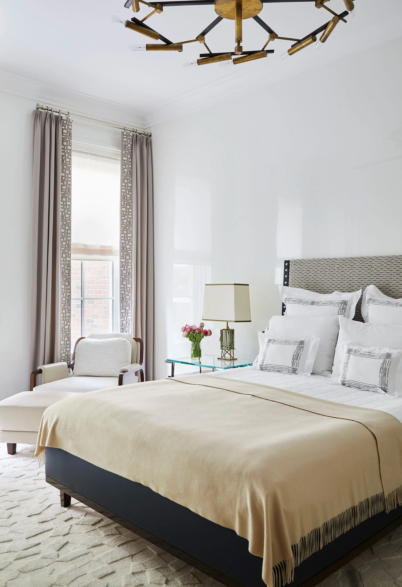 A transitional bedroom featuring a bed with white and beige bedding, a glass nightstand, a cream armchair, and a geometric gold chandelier.