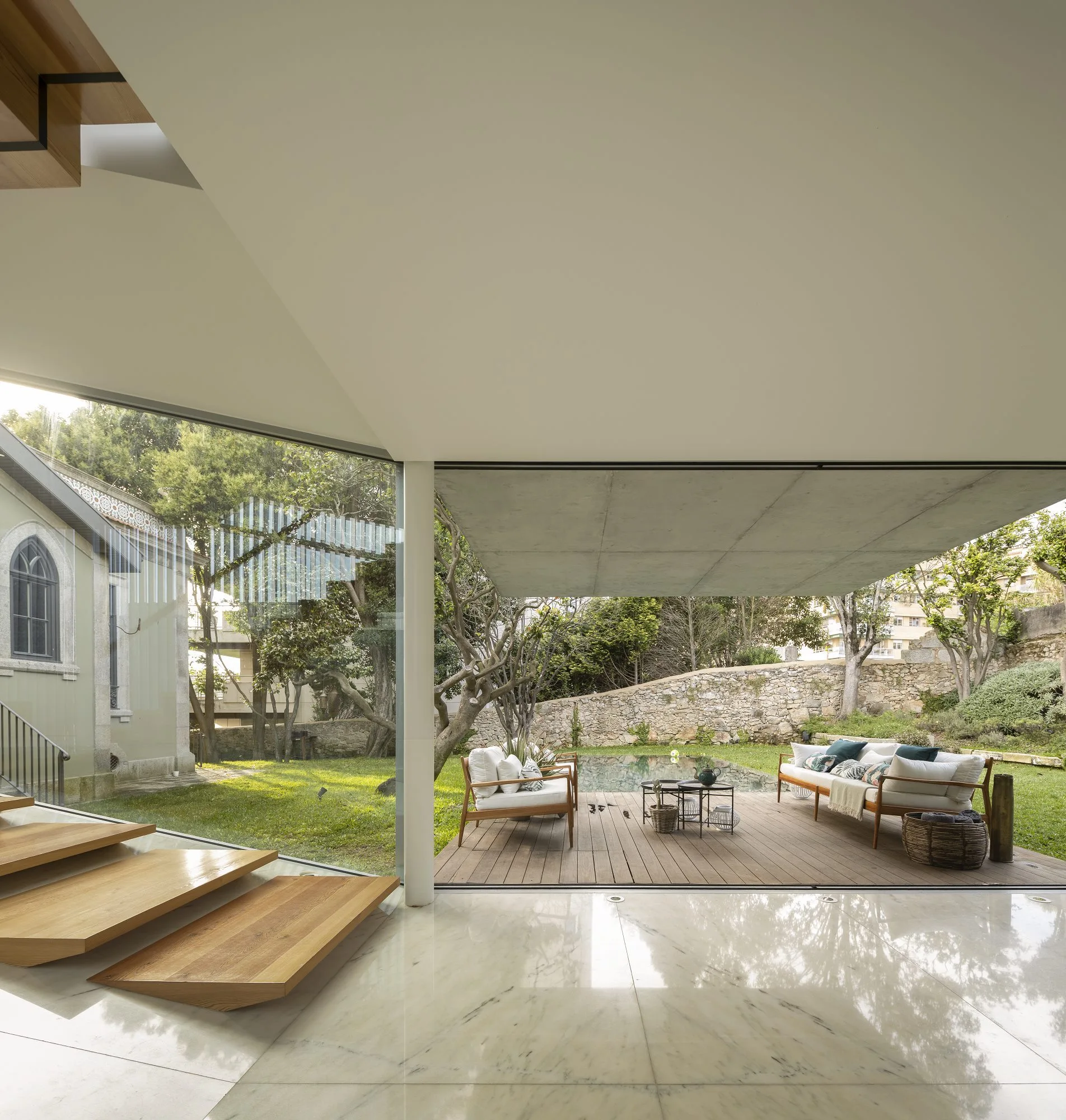 A modern indoor-outdoor space features a polished white floor and floating wooden staircase opening onto a covered patio deck with a sofa and pool.