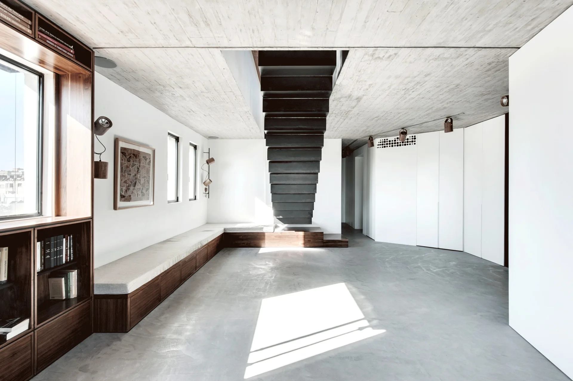 A minimalist room features a dark metal floating staircase, raw concrete ceilings, and a built-in wooden L-shaped bench along white walls.