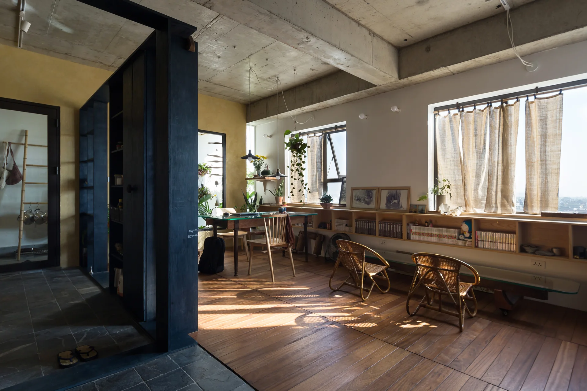 An open dining and work space features wood flooring, a raw concrete ceiling, a low wooden bookshelf under the windows, and a dark room divider.