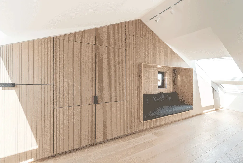 A bright attic room featuring light wood slatted wall cabinets, a recessed seating niche with a dark cushion, and light wood flooring.