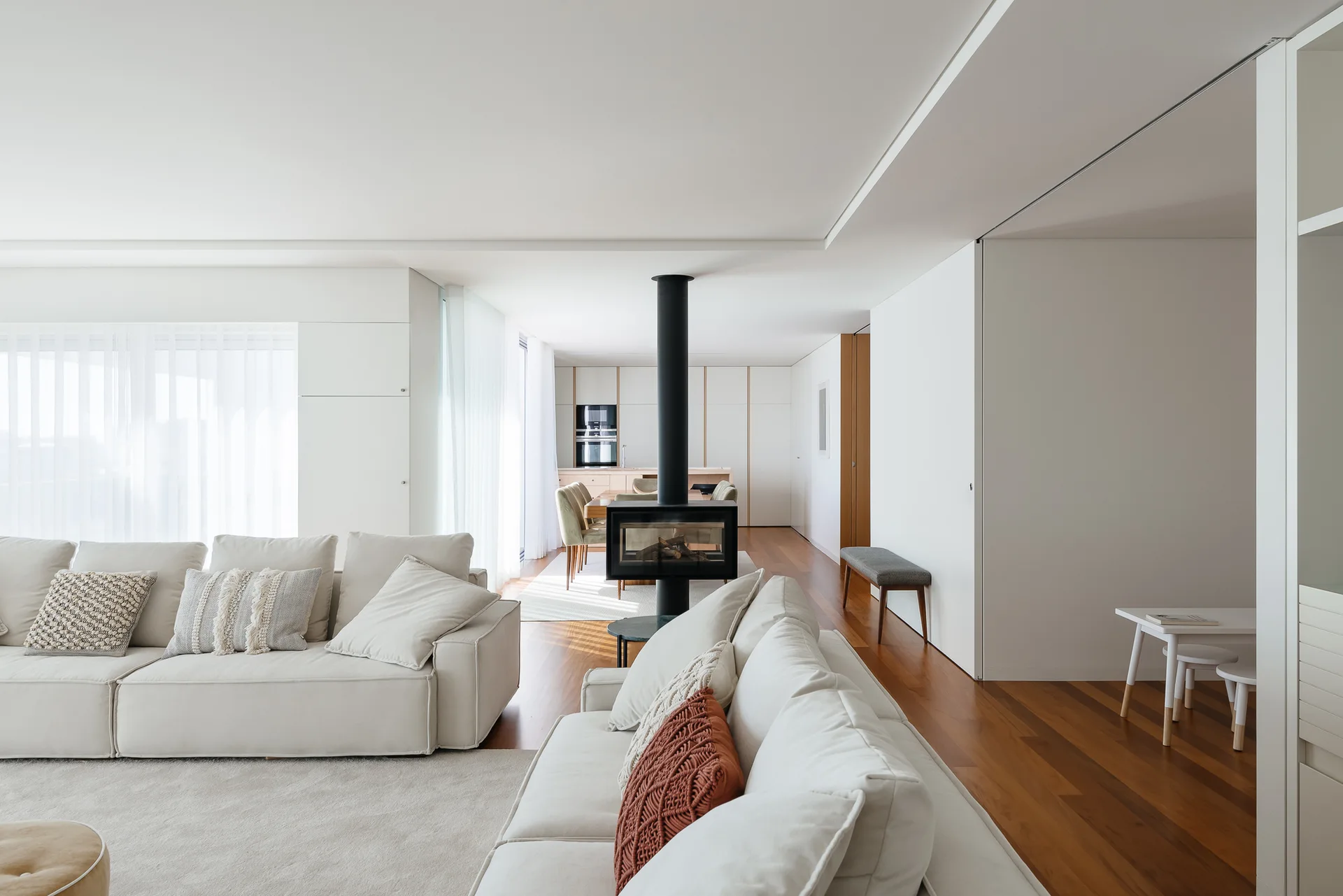 A modern open-concept living room features off-white sofas, warm wood flooring, and a freestanding black fireplace column.