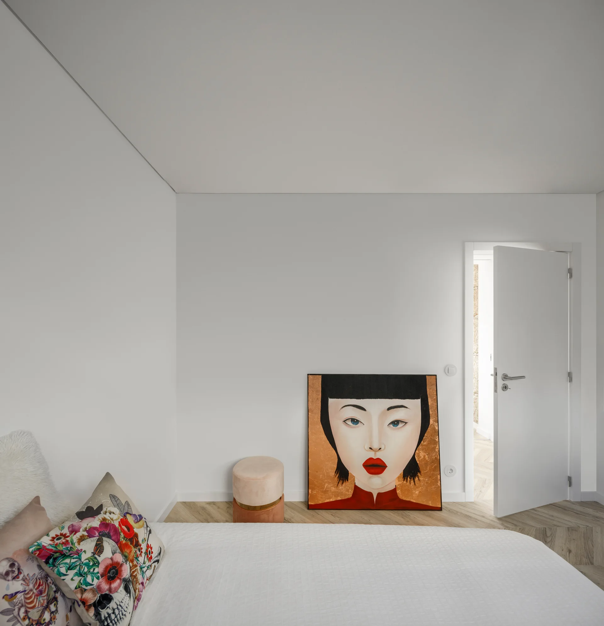 A minimalist white bedroom featuring a colorful portrait painting leaning on the wood floor near a bed with floral throw pillows.
