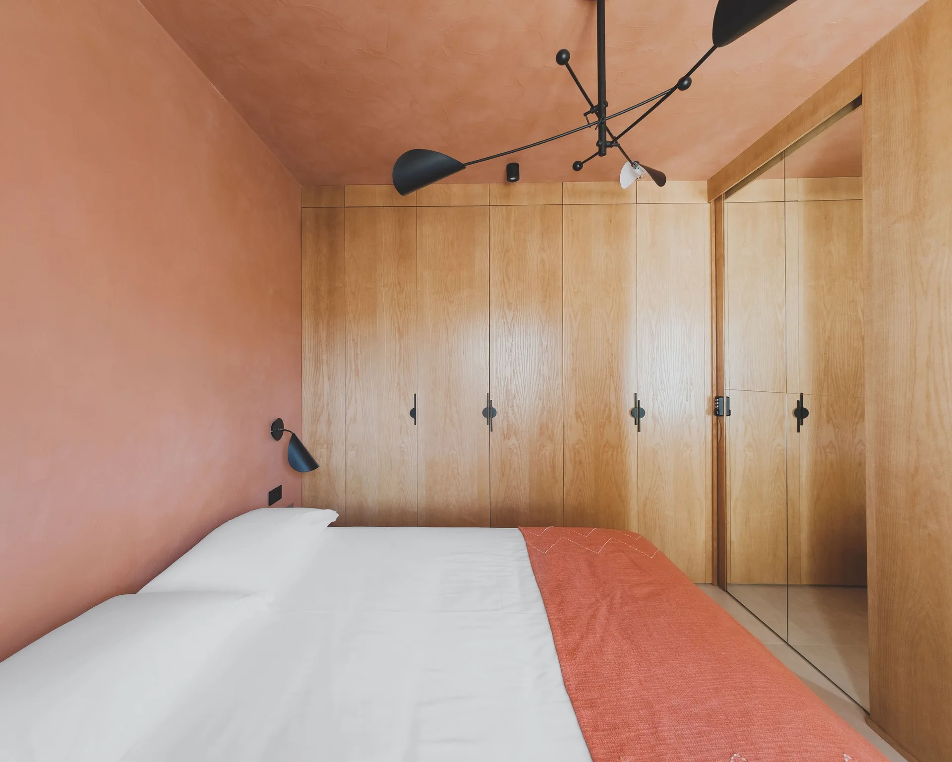A contemporary bedroom features terracotta plaster walls and ceiling, a bed with white linens, and a full wall of built-in wooden wardrobes.