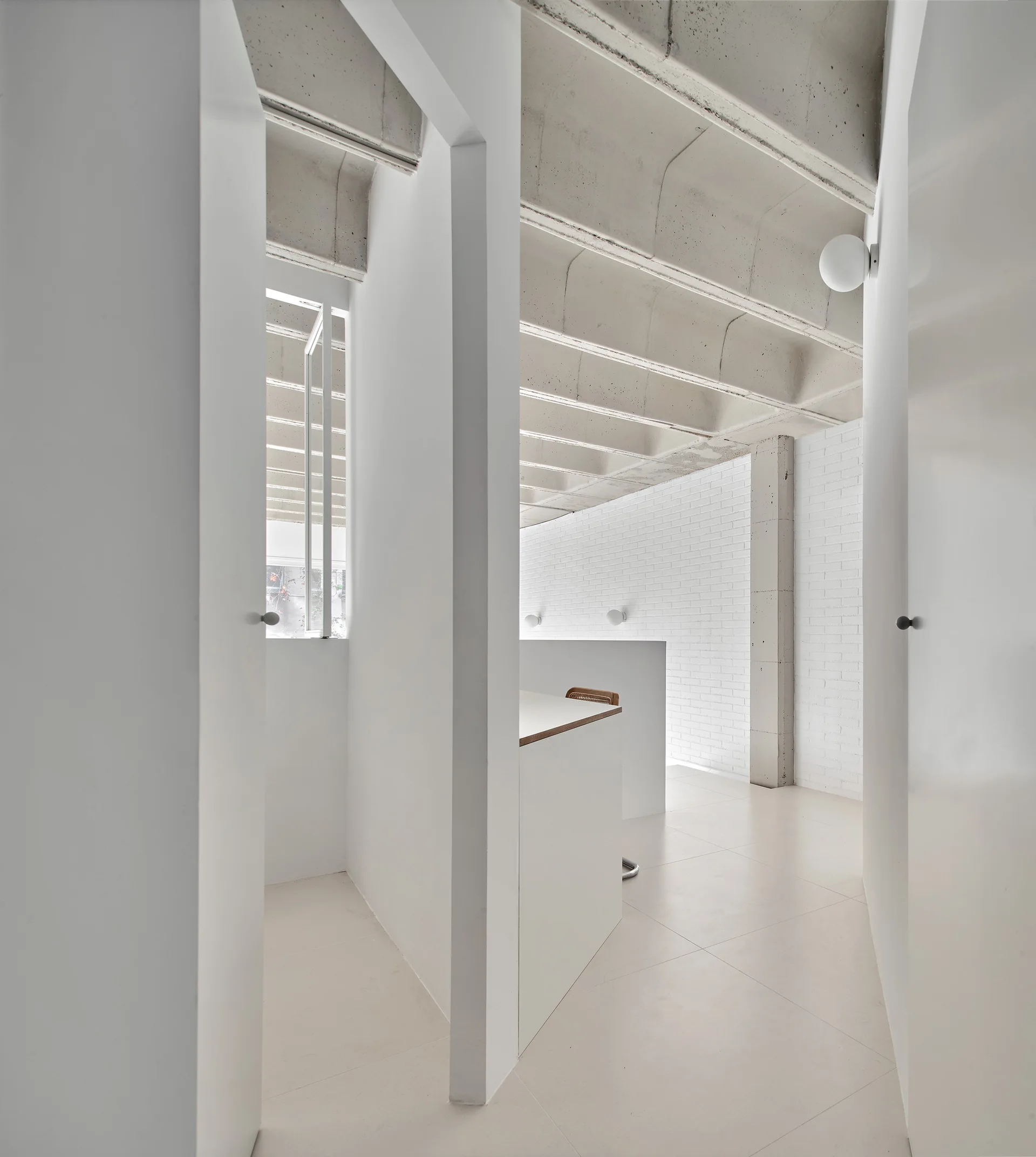 A minimalist white hallway features an exposed concrete ribbed ceiling, smooth partition walls, and a painted brick wall with globe sconces.