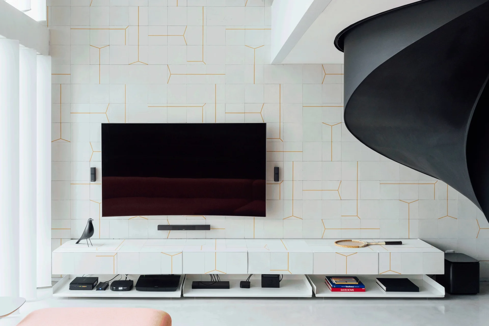 A minimalist living room featuring a wall-mounted TV on white geometric tiles, a matching low console, and a black spiral staircase.