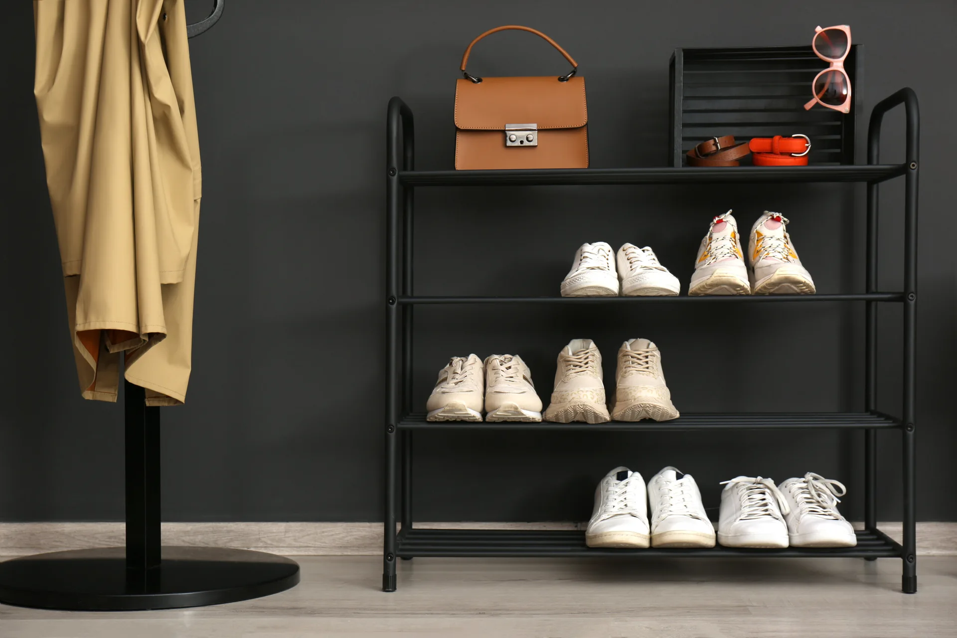 A four-tier black metal shoe rack displaying sneakers and a leather handbag stands against a dark grey wall.