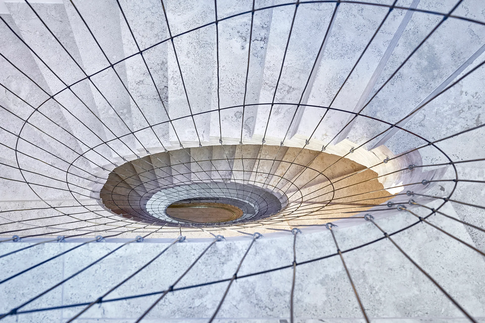 A top-down view looking down a spiraling stone staircase fitted with a taut black wire mesh safety railing.
