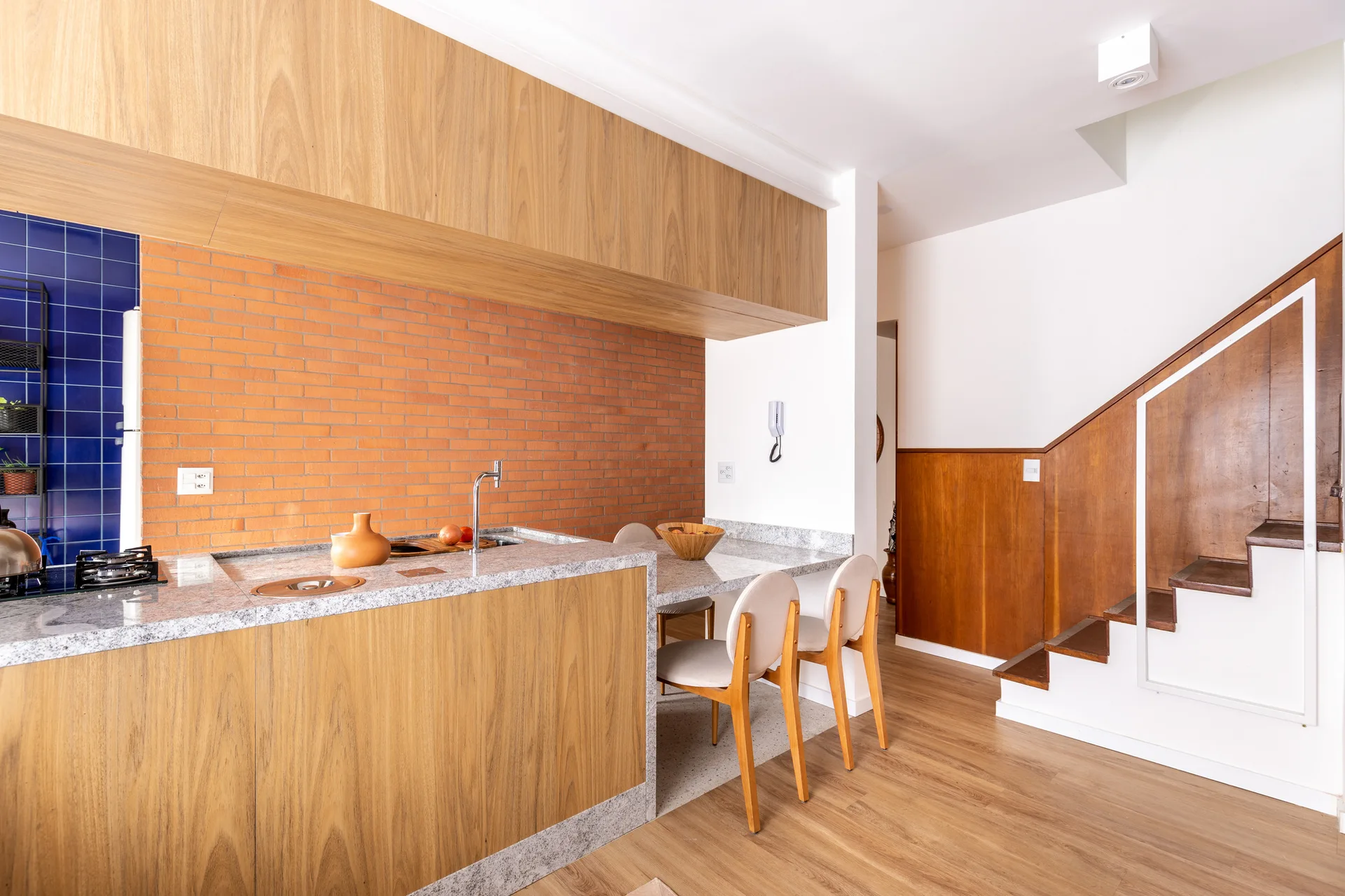 A residential kitchen featuring a wood-wrapped island with granite countertops, an exposed brick wall, and a wooden staircase on the right.