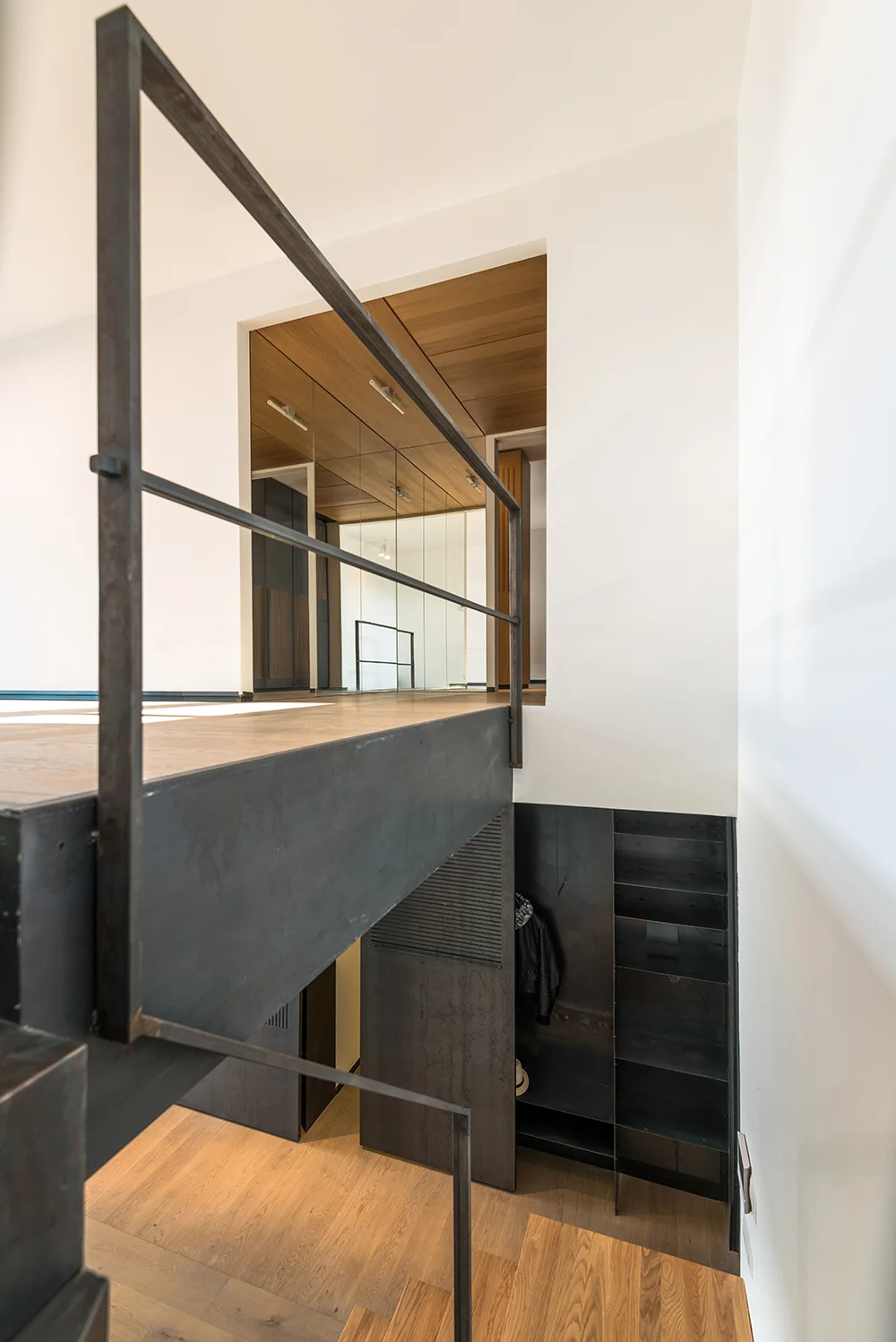 A minimalist home interior features a steel staircase with industrial railings, warm wood-paneled ceilings, and dark built-in metal shelving.