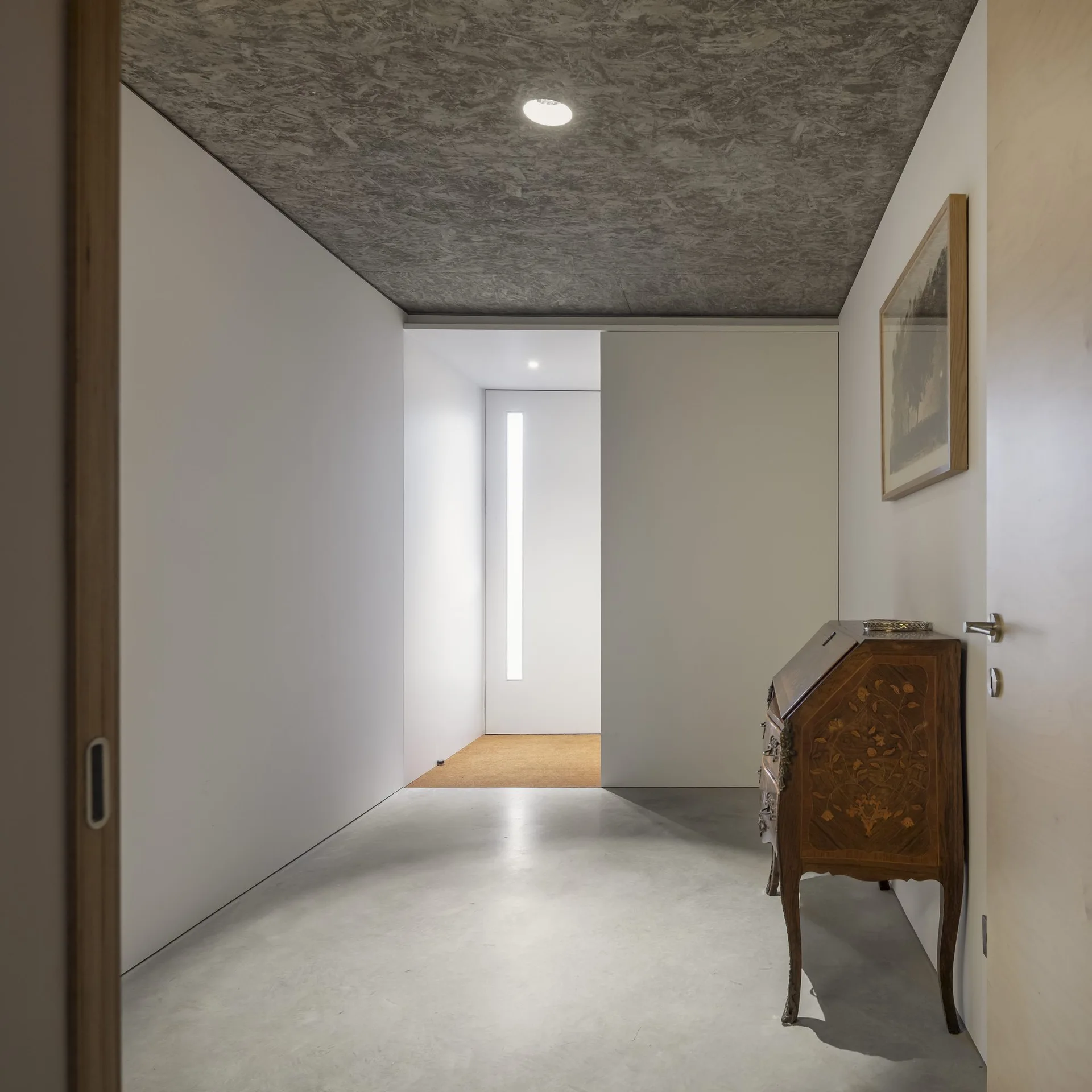 A modern minimalist hallway with grey concrete flooring, white walls, a textured OSB ceiling, and an antique wooden bureau.