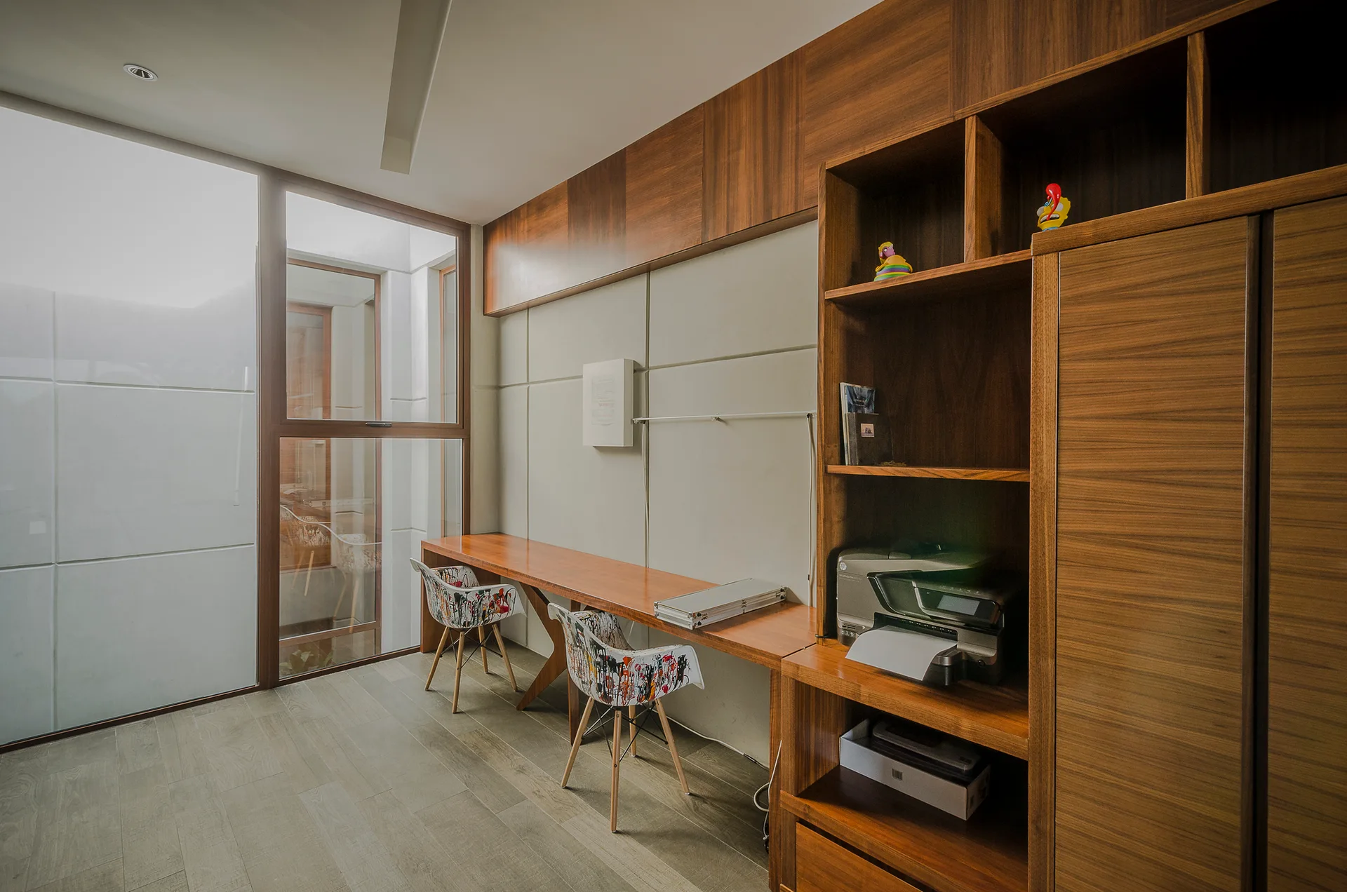 A modern home office featuring a long built-in wooden desk, walnut shelving, two patterned chairs, and large white wall panels beside a tall window.