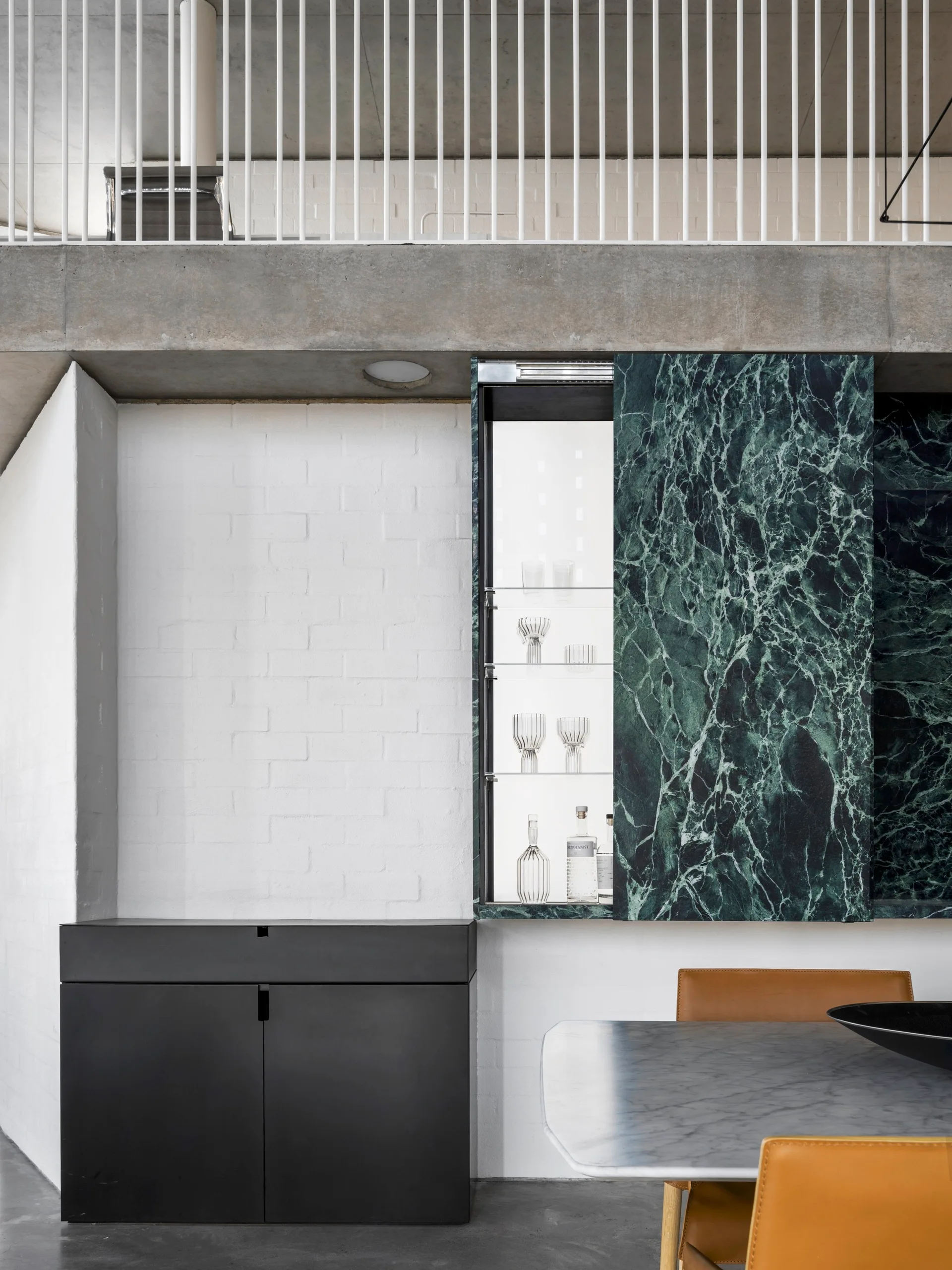 A modern dining area features a recessed bar cabinet with a sliding green marble panel, set against a painted white brick wall.
