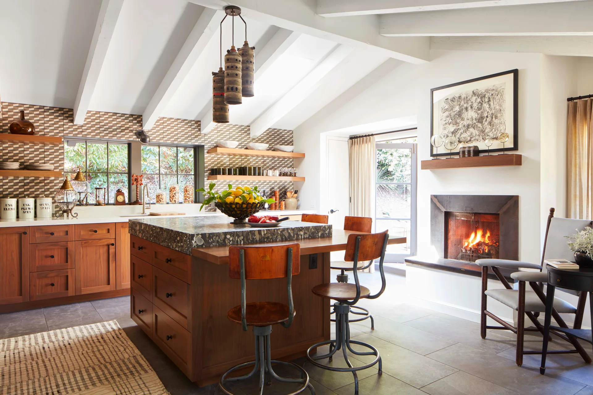 A rustic kitchen features a wooden island with vintage swivel stools, a geometric tile backsplash, and a burning fireplace in the wall.