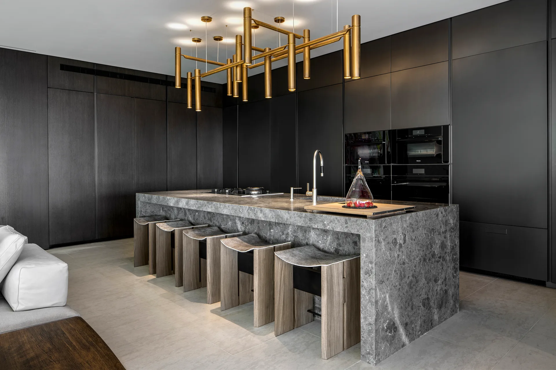 A gray stone kitchen island with curved wooden stools sits beneath a geometric gold pendant light in front of matte black cabinetry.