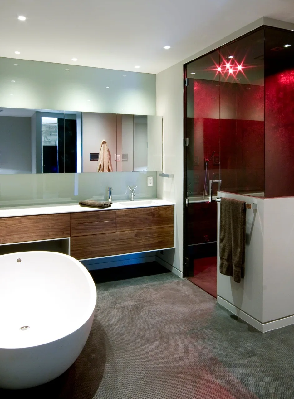 A modern bathroom features a white freestanding bathtub, a floating wood vanity with integrated sinks, and a glass shower enclosure with red walls.