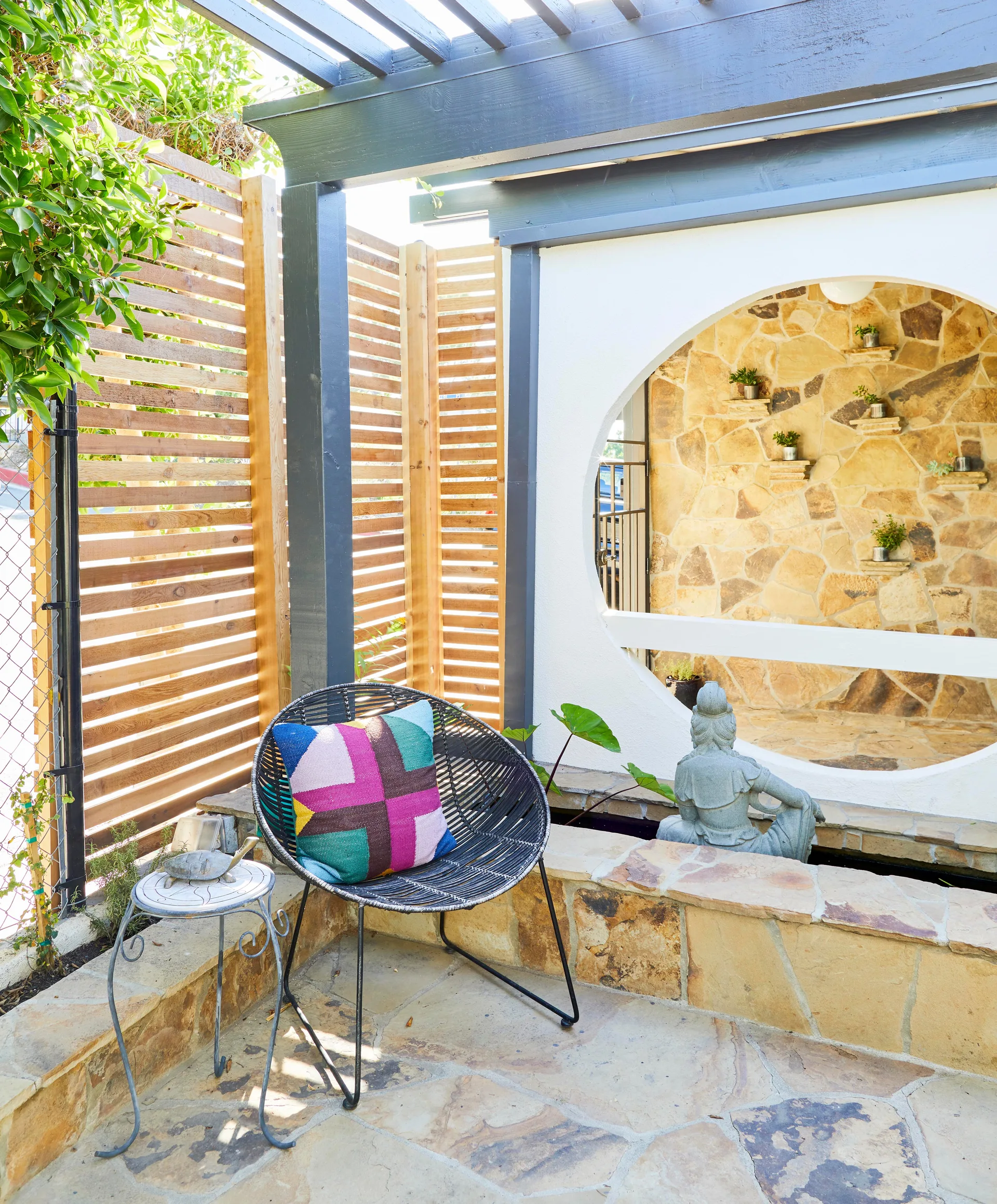 A flagstone patio features a black woven lounge chair, a horizontal wood slat privacy screen, and a wall with a circular cutout revealing a stone wall.