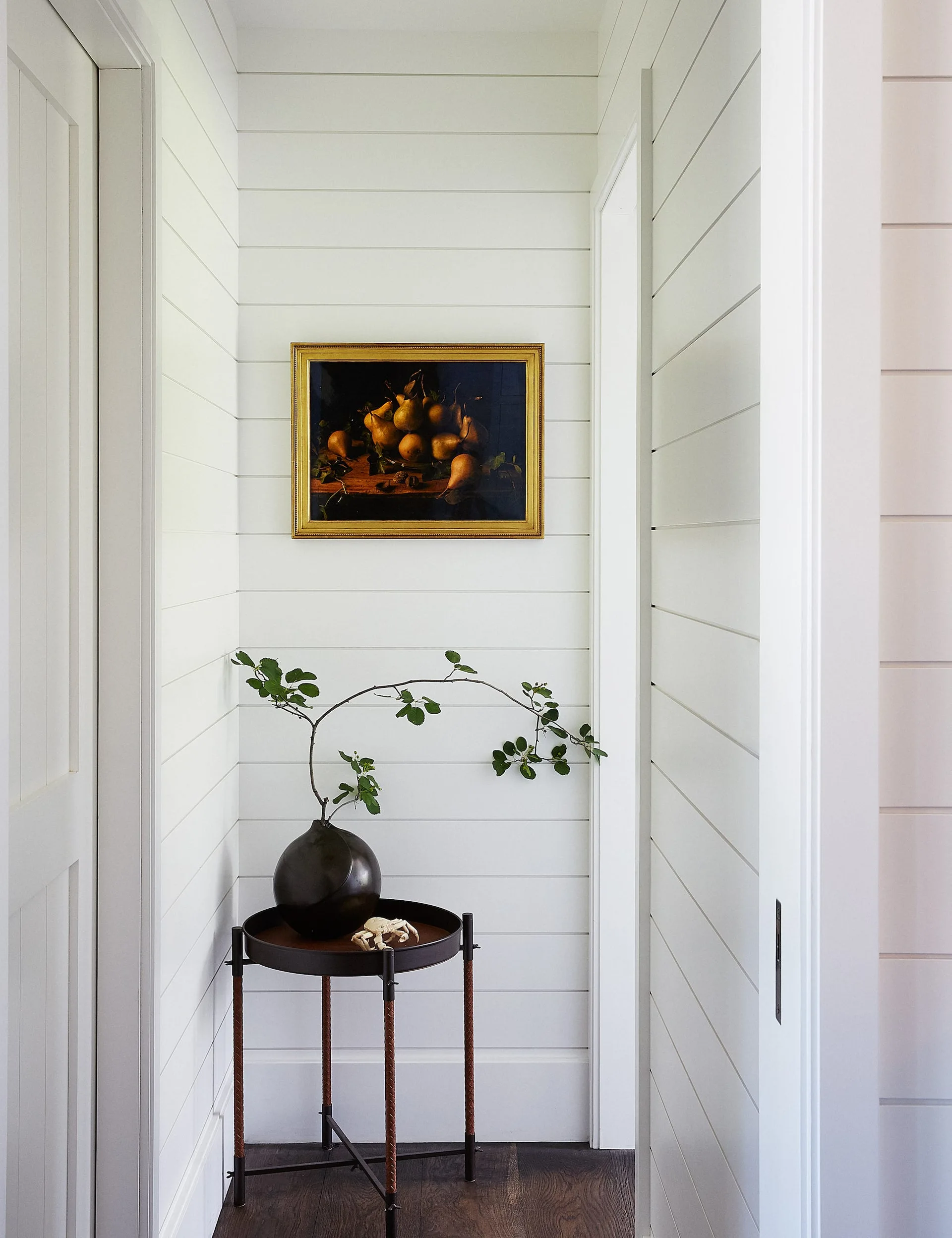 A small round accent table with a green branch in a black vase sits below a gold-framed still-life painting on white shiplap hallway walls.