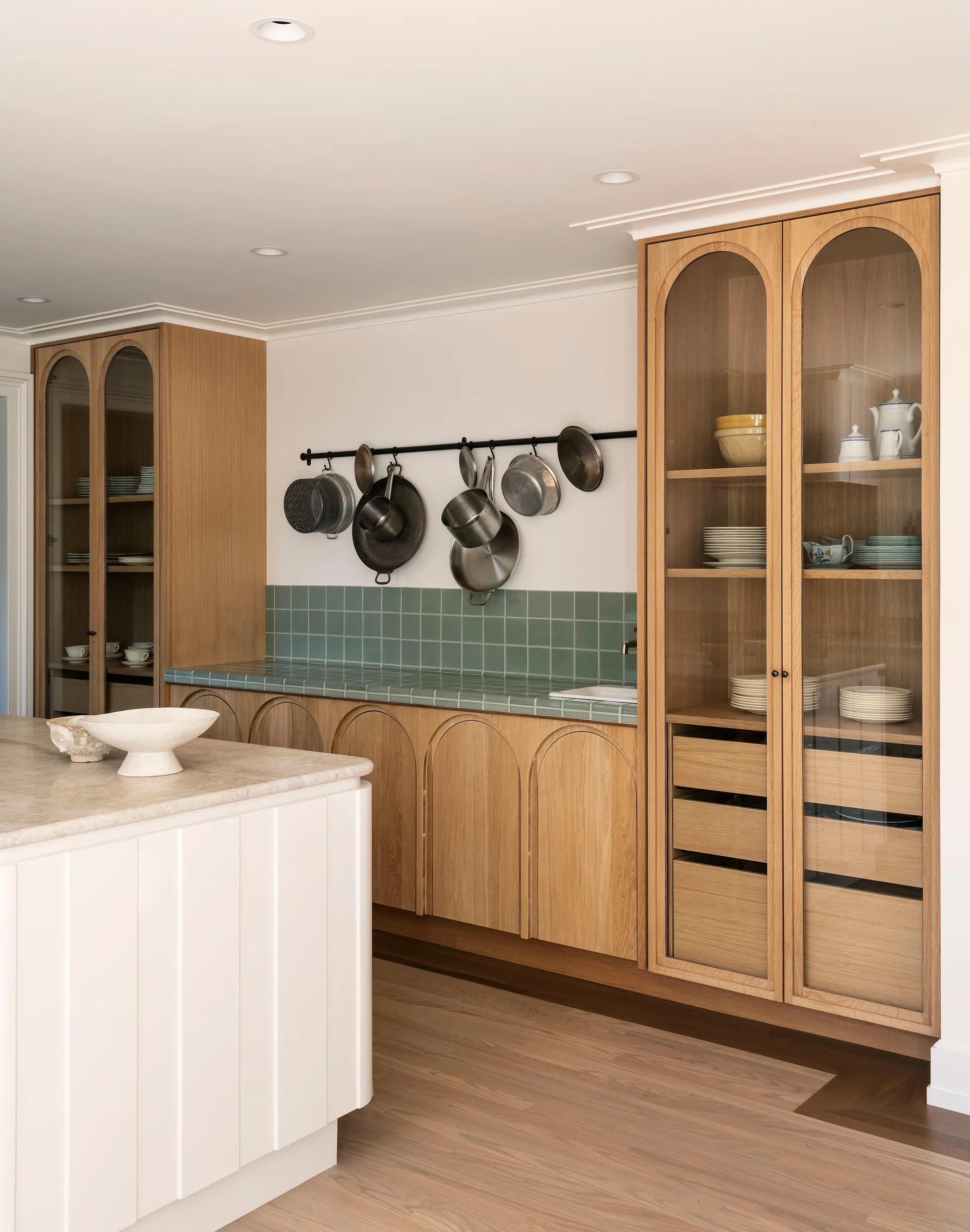 A kitchen features tall light oak cabinets with arched glass doors, a green tile backsplash and countertop, and a white paneled island.