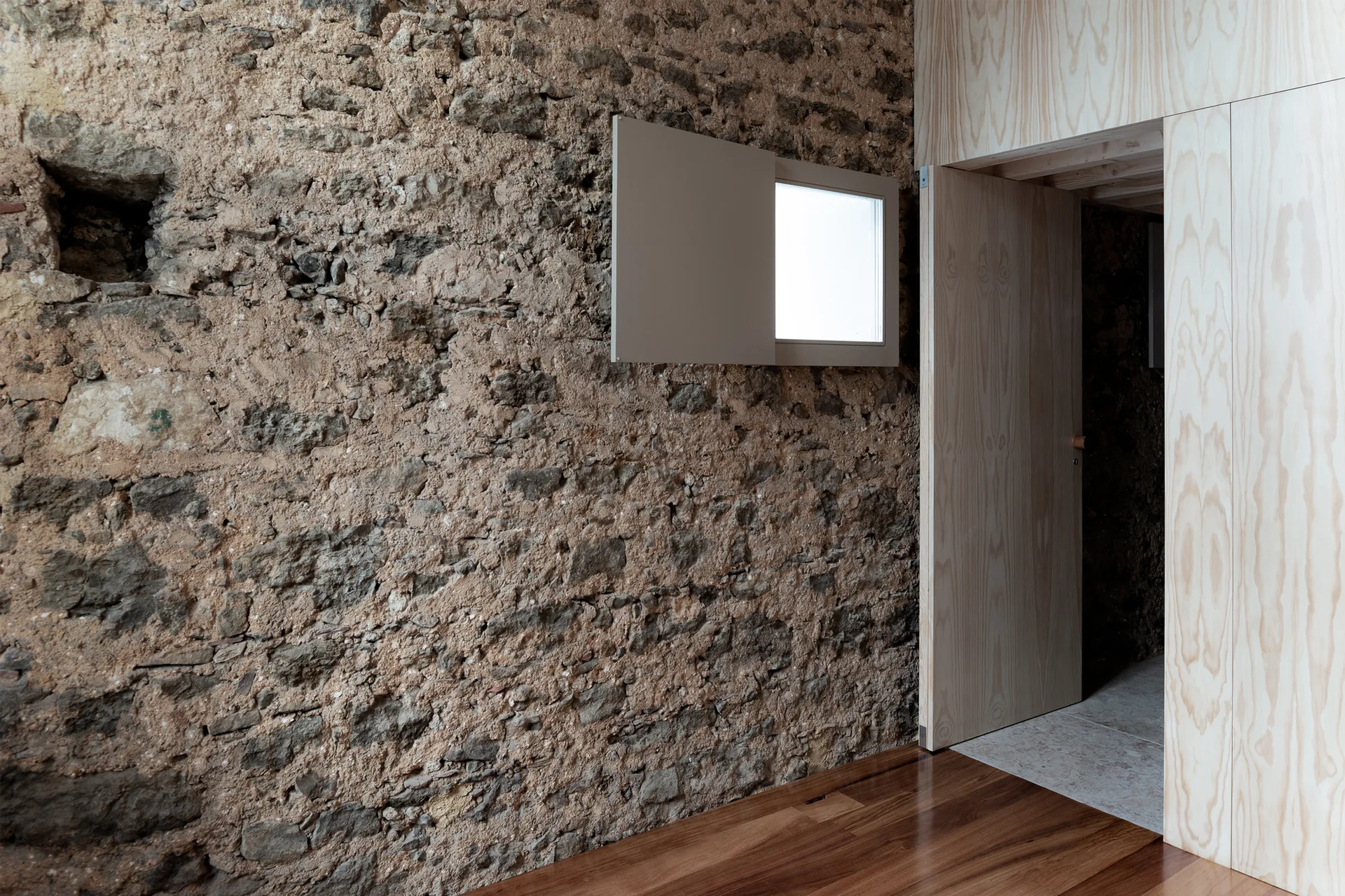 An interior view of a rough exposed stone wall with a small window next to a smooth plywood partition and open doorway.