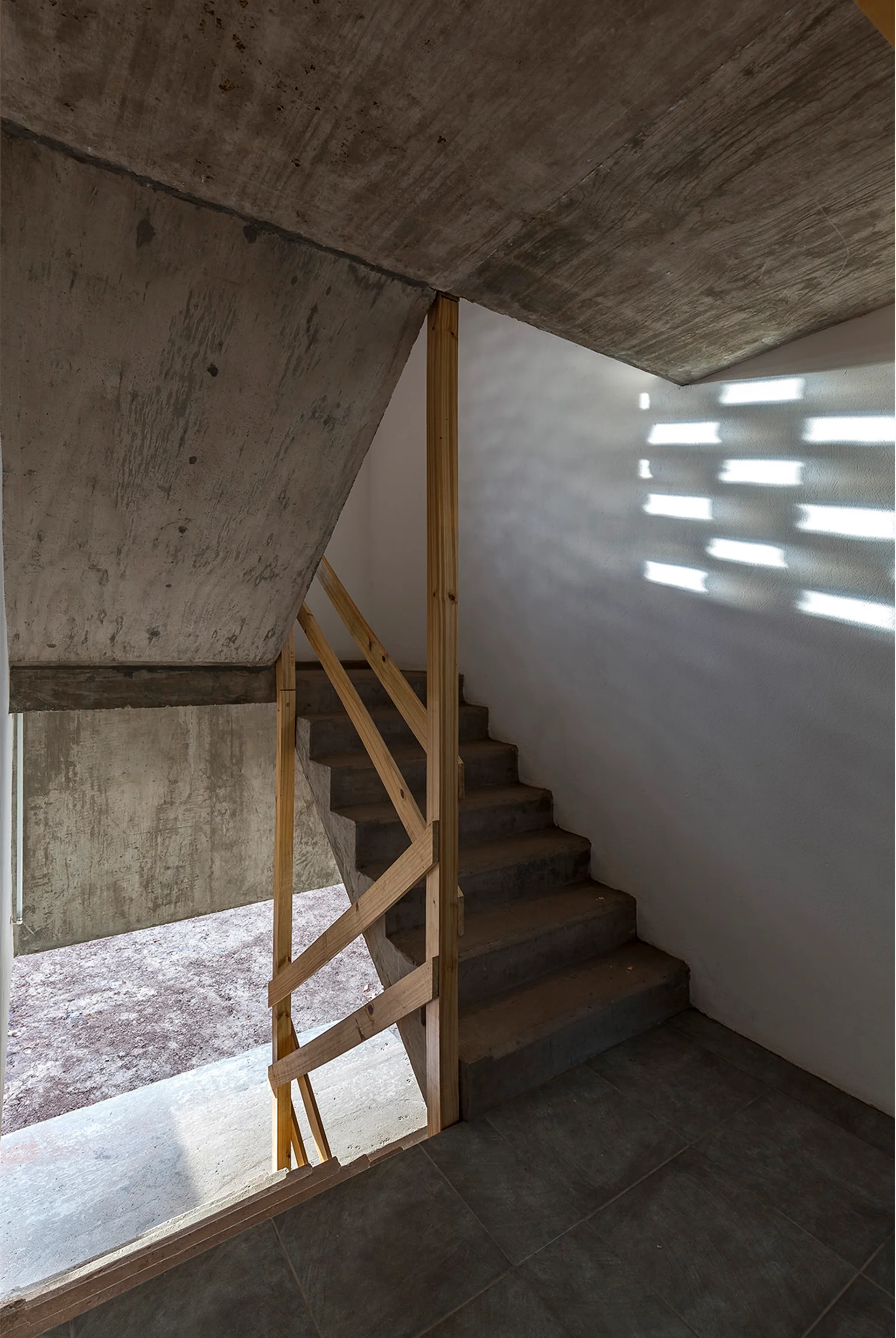 An interior concrete staircase featuring a simple wooden timber railing against white plaster walls with geometric sunlight patterns.