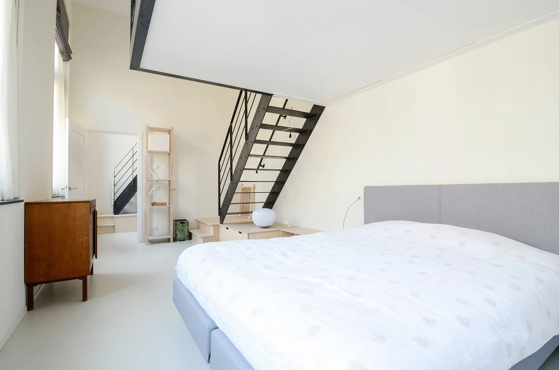 A minimalist bedroom featuring a grey upholstered platform bed, a black metal staircase, white painted floors, and a vintage wooden cabinet.