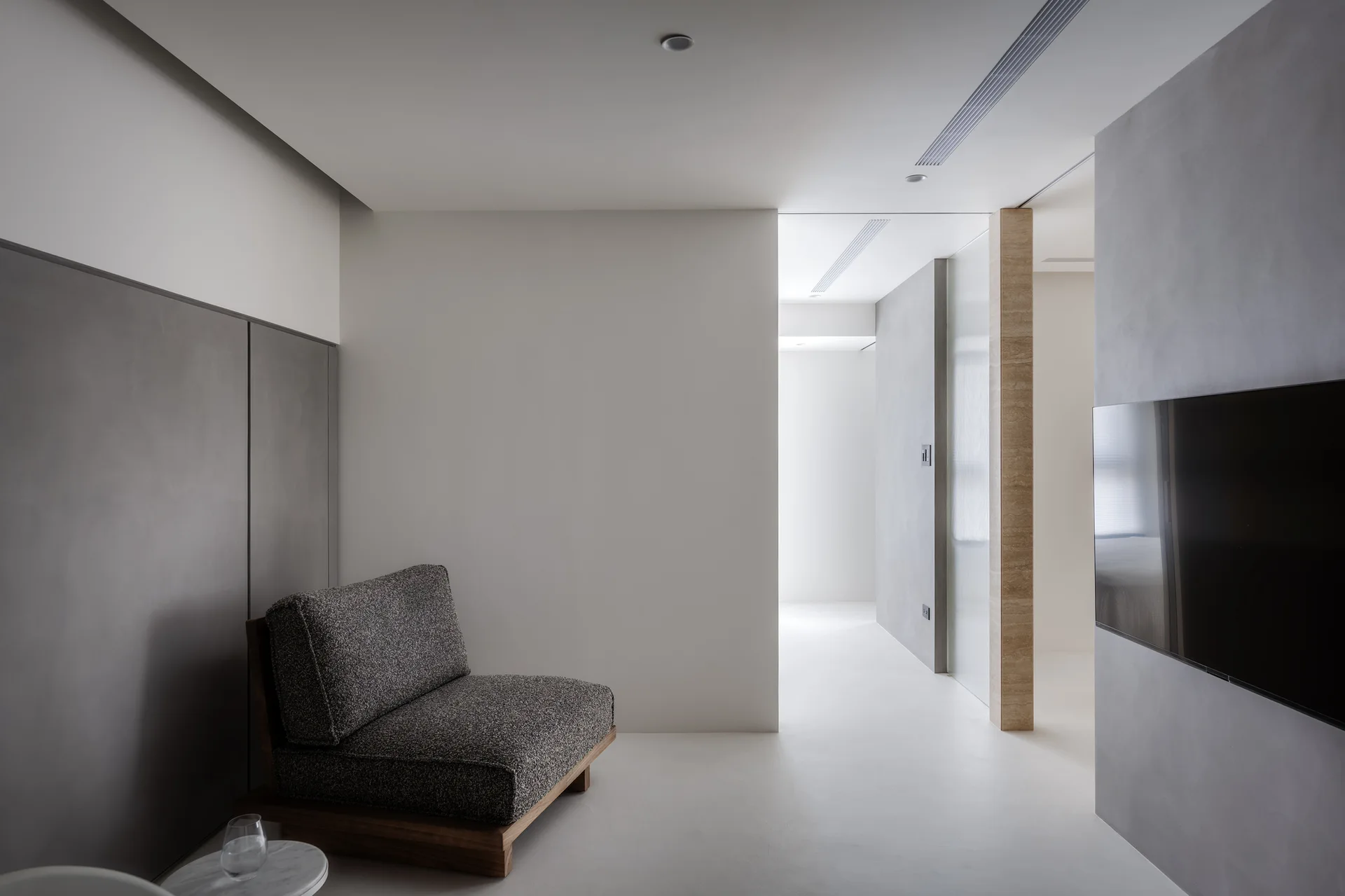 A minimalist room featuring a low-profile textured grey armchair next to smooth grey wall panels, with a beige stone pillar in the background.