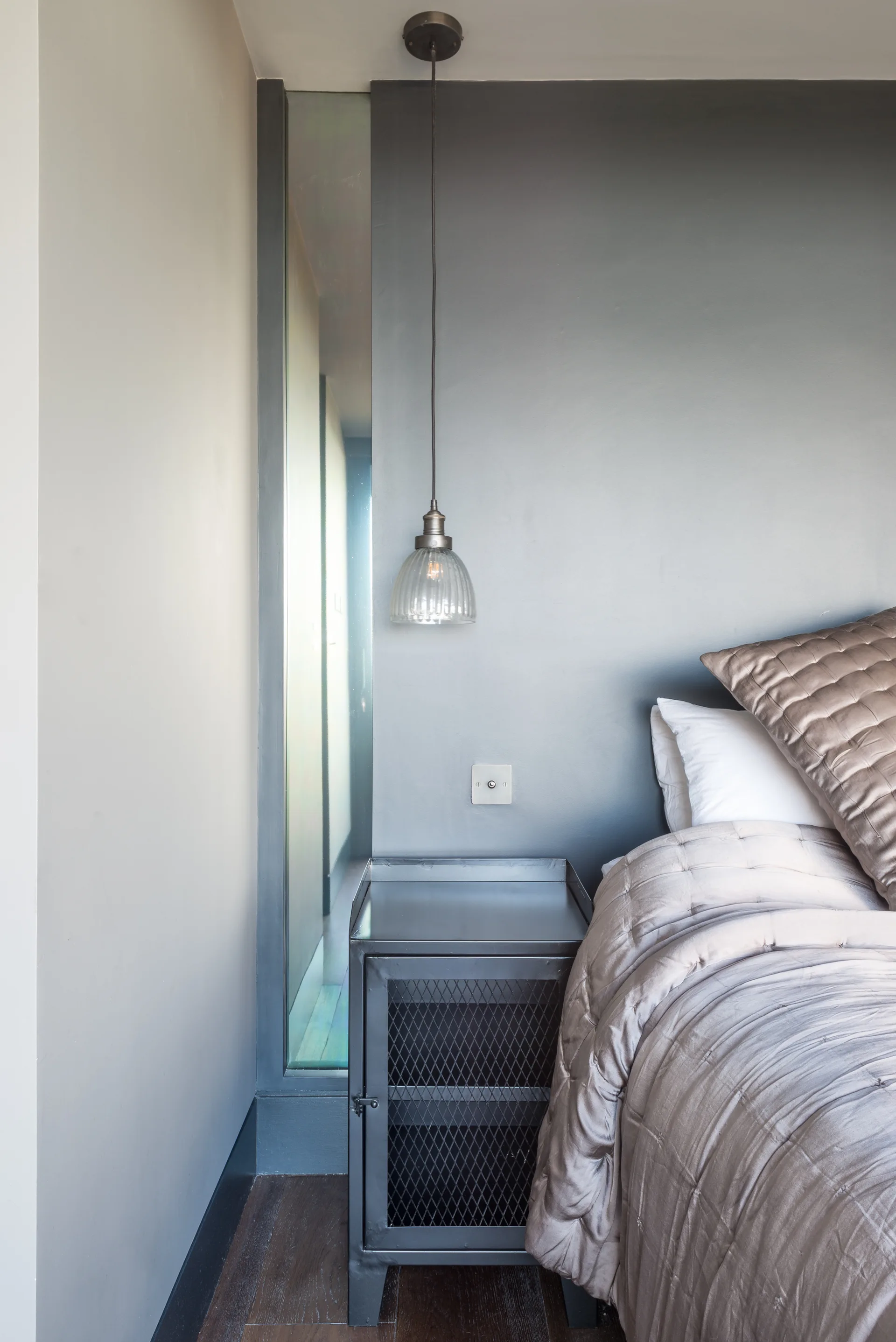 A bedroom corner features a grey quilted bed beside an industrial metal mesh nightstand under a ribbed glass pendant light hanging from the ceiling.