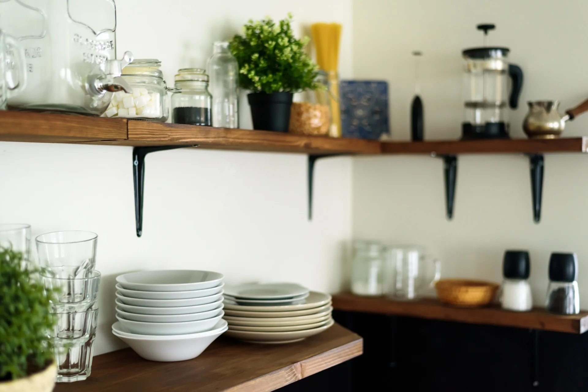 Minimalist open wood shelves on an off-white wall display stacked white bowls, clear drinking glasses, and glass pantry jars.