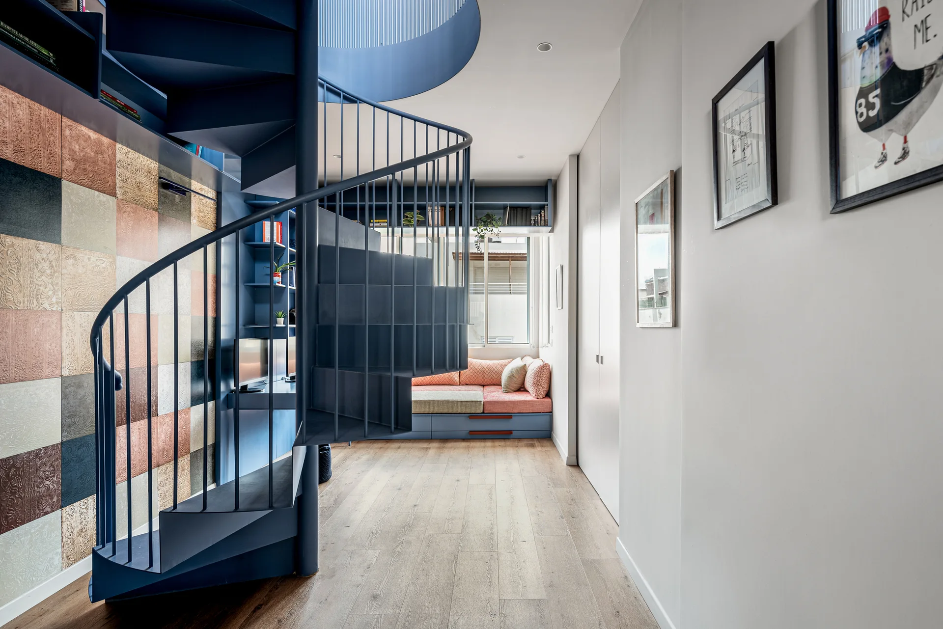 A blue metal spiral staircase stands in a modern hallway next to textured wall panels and a built-in daybed beneath a window.