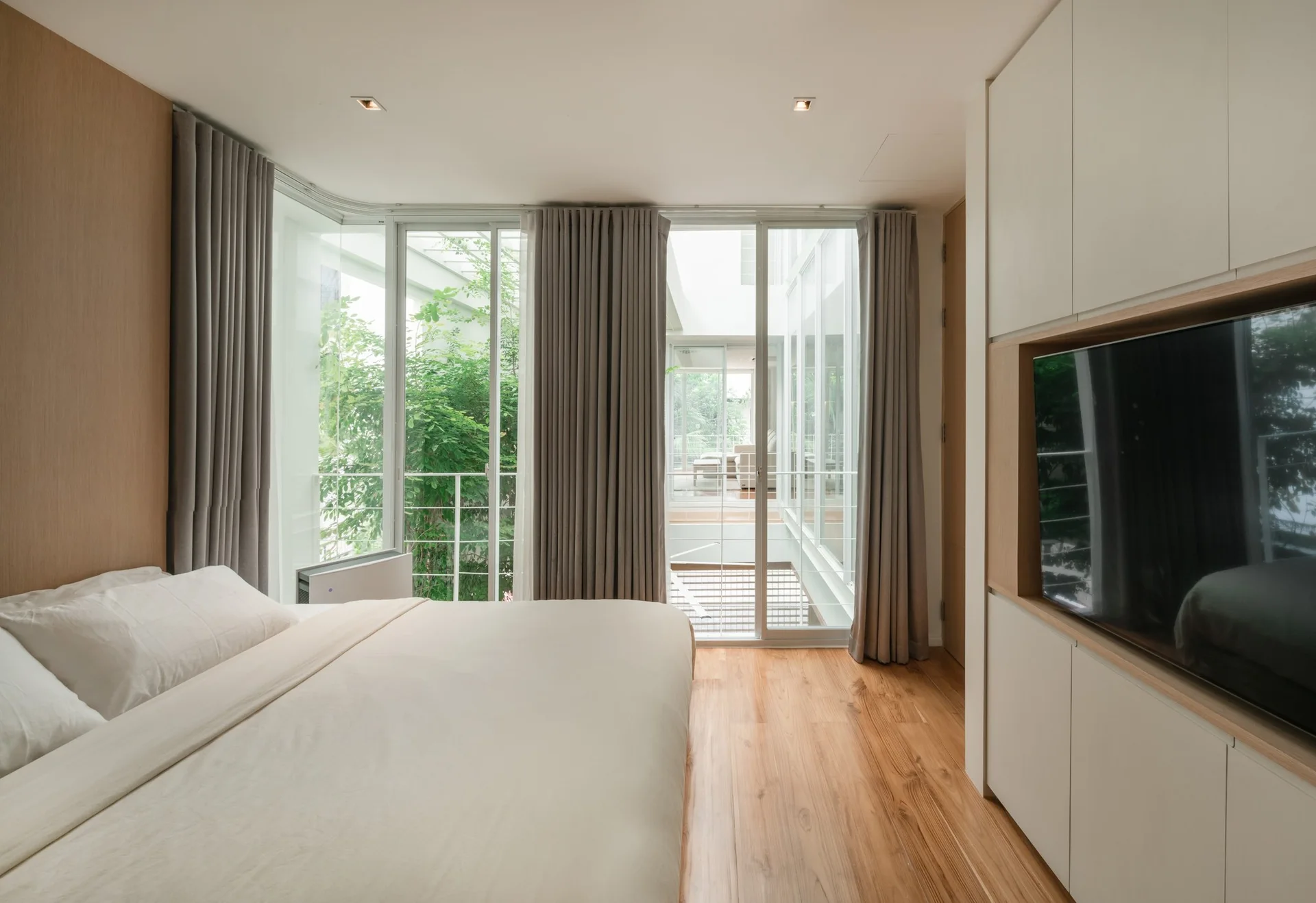 A minimalist bedroom features a bed with white bedding, wood floors, a built-in white media cabinet with a television, and large glass sliding doors.
