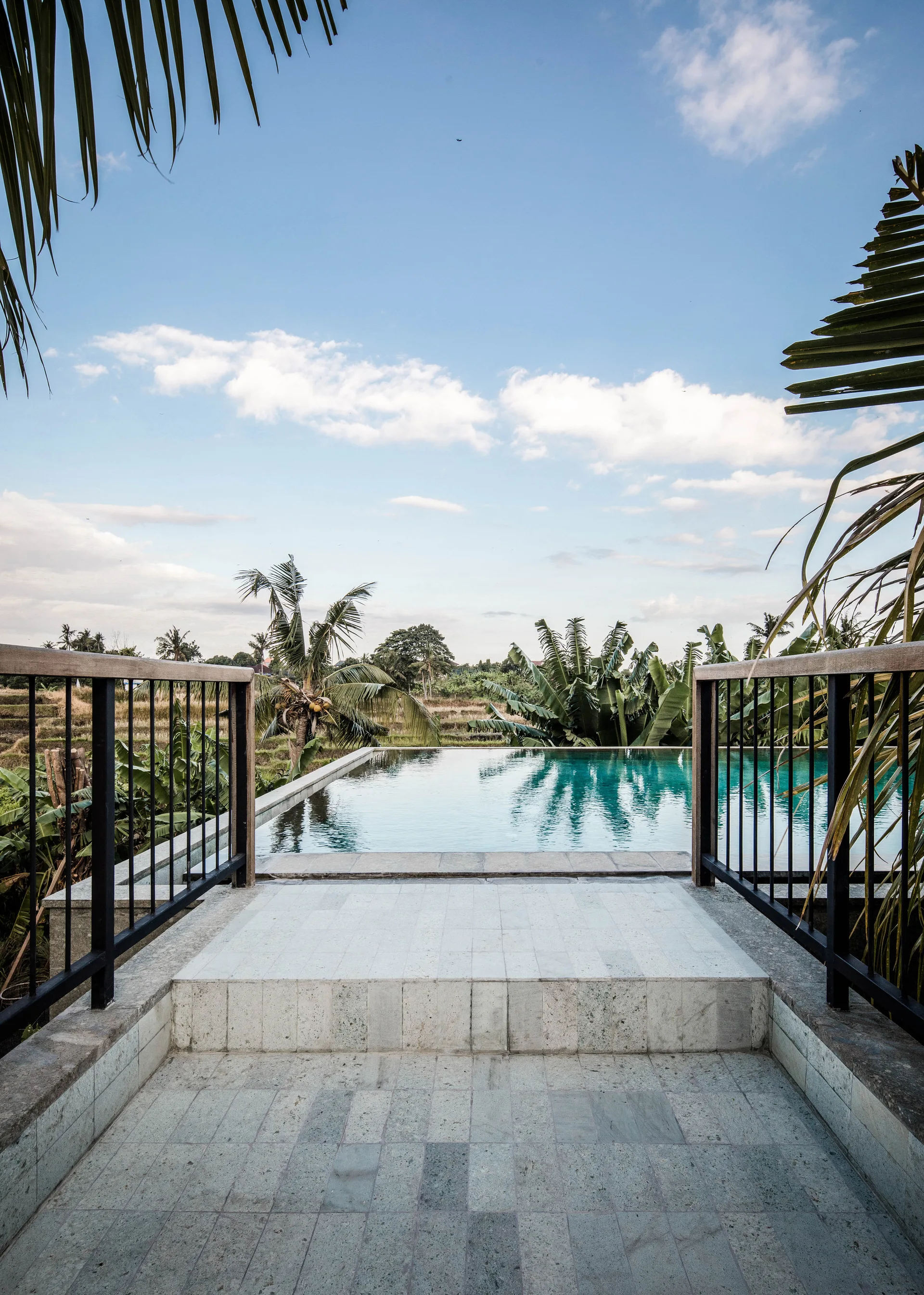 A stone paved walkway with black metal railings leads directly to a rectangular infinity pool overlooking a tropical green landscape.