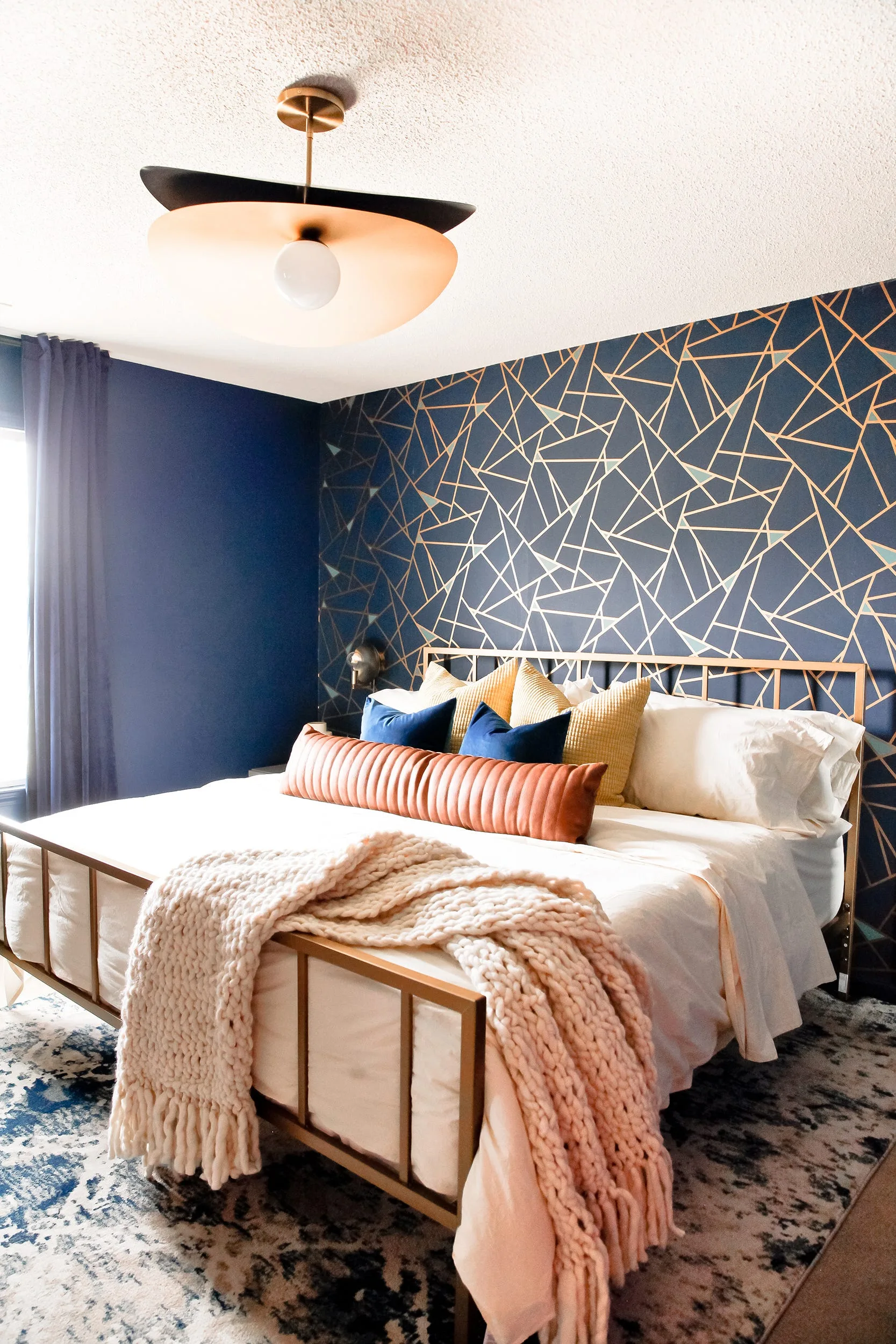 A modern bedroom features a brass bed frame with colorful pillows against a navy blue geometric wallpapered accent wall.