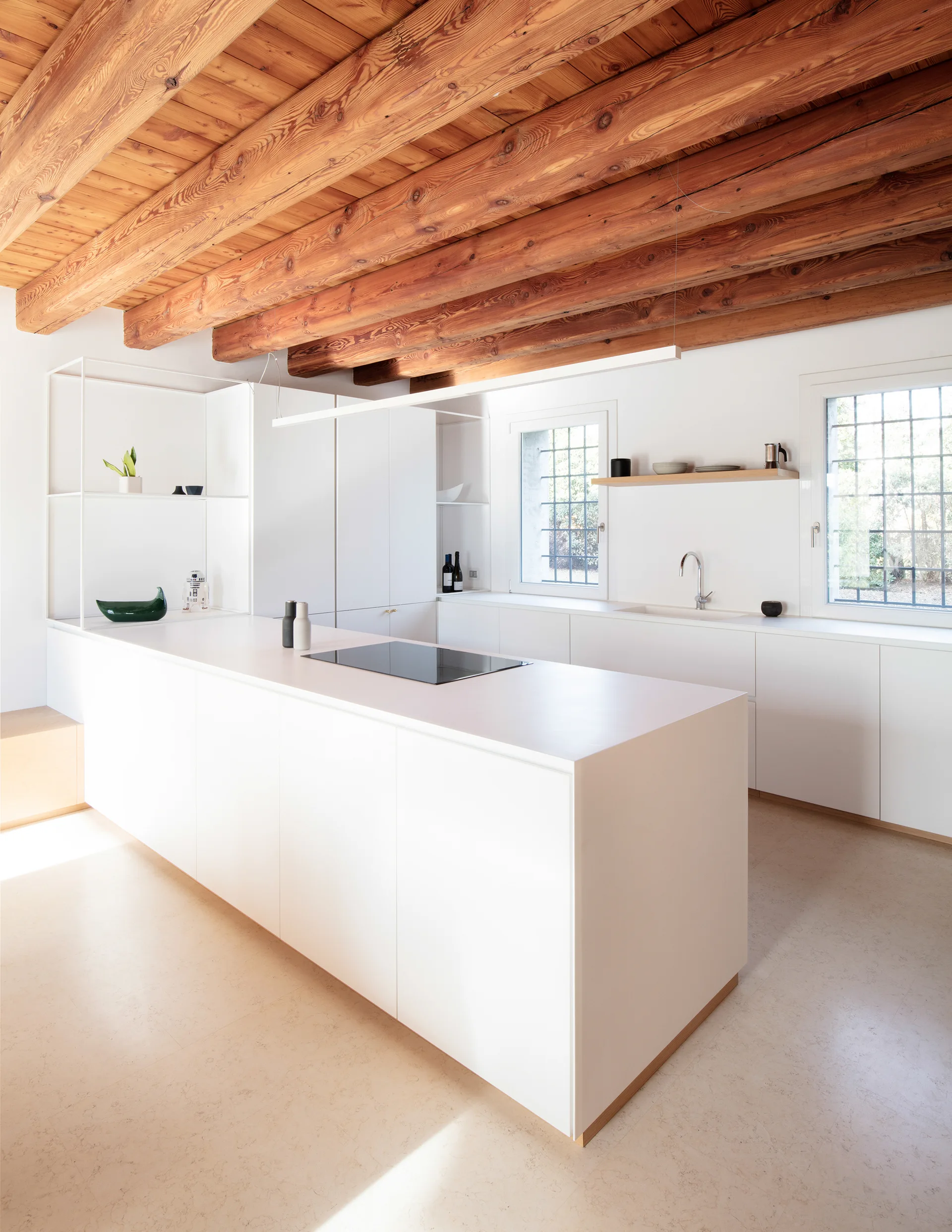 A minimalist kitchen features a white central island, handleless cabinetry, and a prominent rustic wood beam ceiling with white-trimmed grid windows.