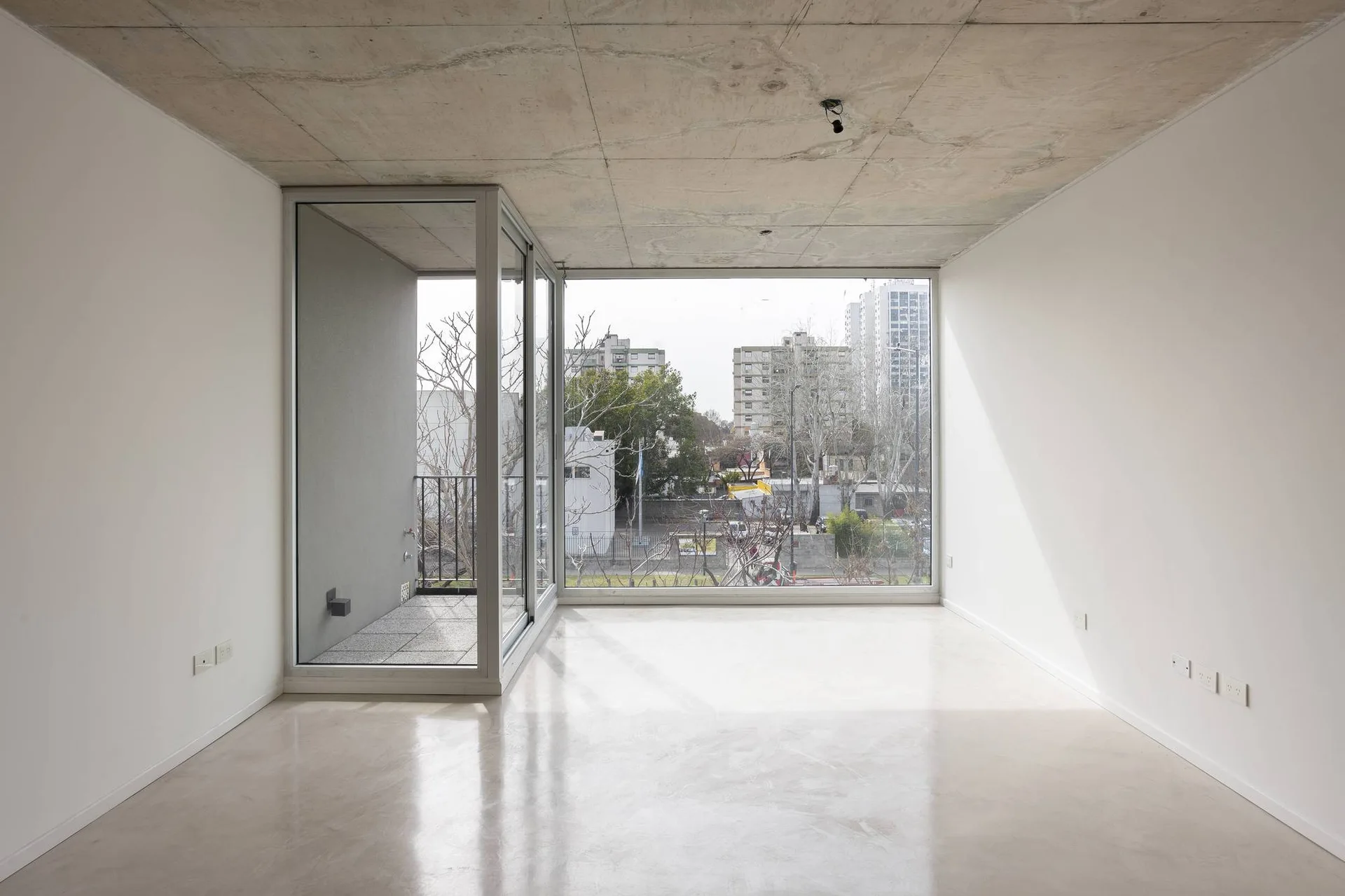An empty minimalist room featuring an exposed concrete ceiling, white walls, polished concrete floors, and a glass sliding door leading to a balcony.