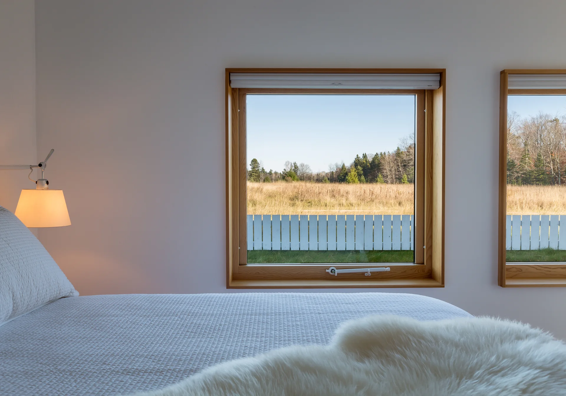 A minimalist bedroom features a bed with quilted white bedding, a sheepskin throw, and large natural wood-framed windows overlooking a rural landscape.