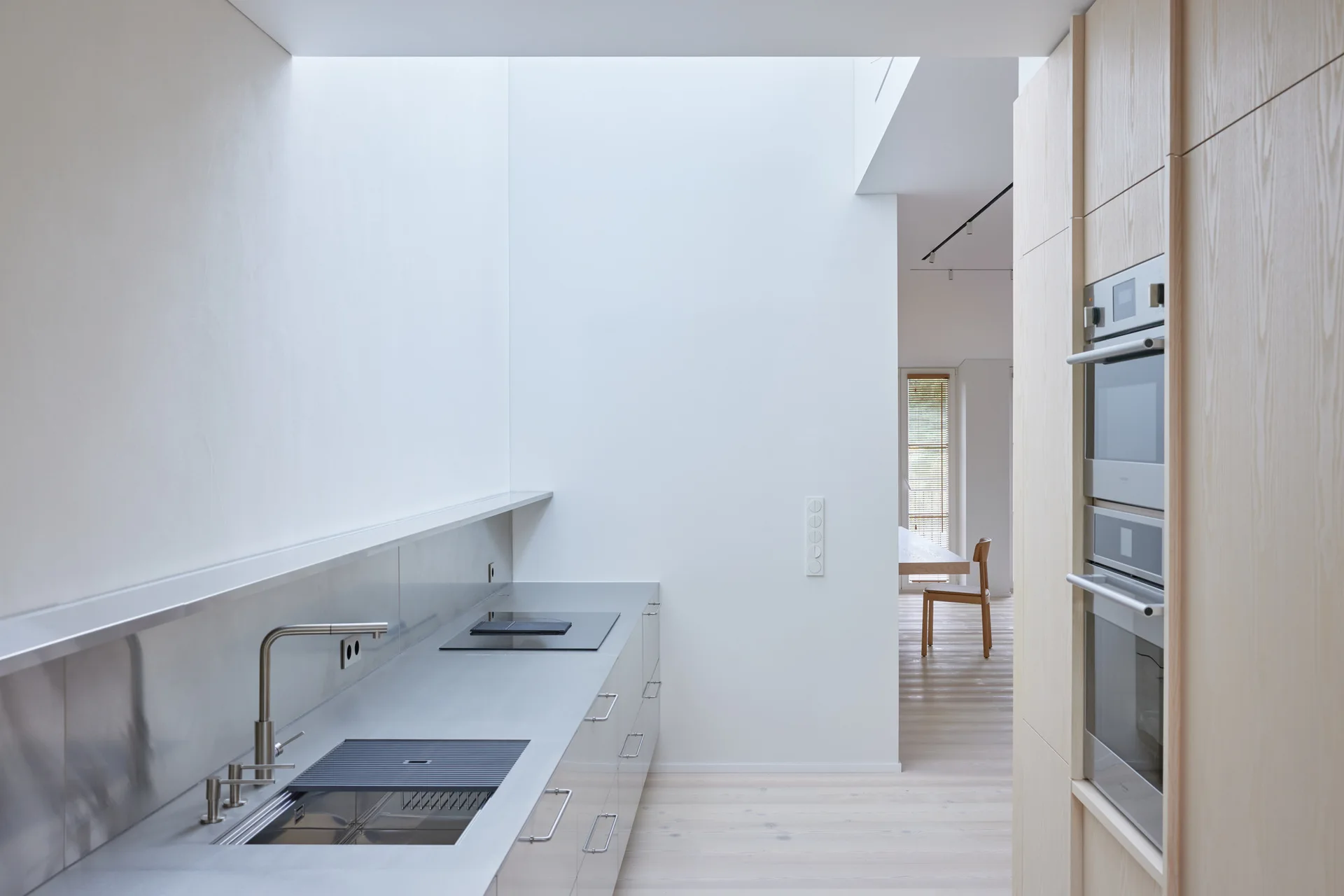 A minimalist kitchen features a stainless steel countertop with a sink, built-in double ovens in wood cabinetry, and a bright overhead skylight.