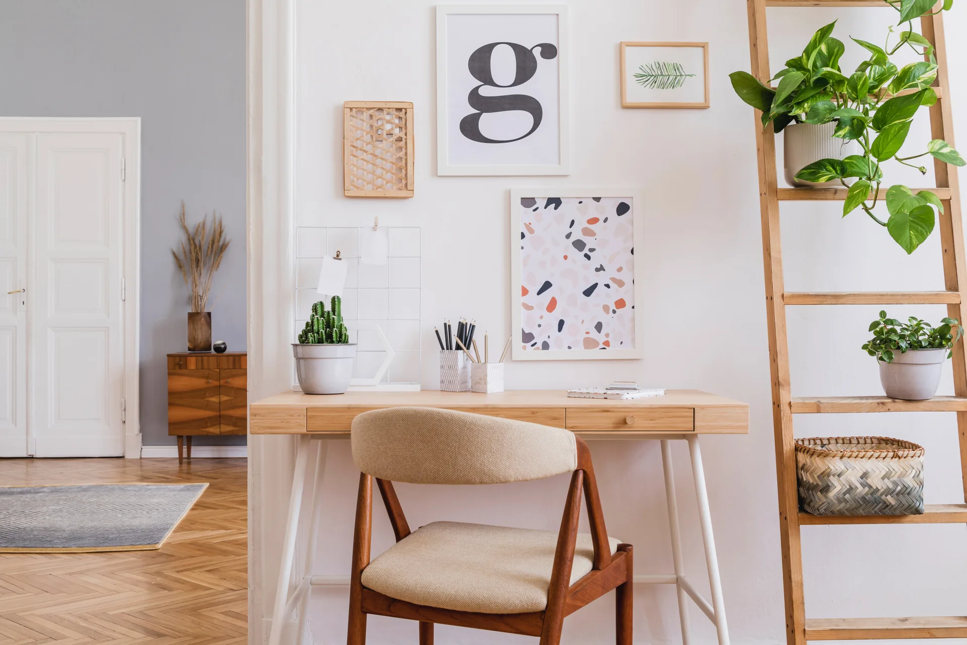 A minimalist workspace features a light wood desk, an upholstered mid-century wooden chair, and a leaning wooden ladder shelf with potted plants.