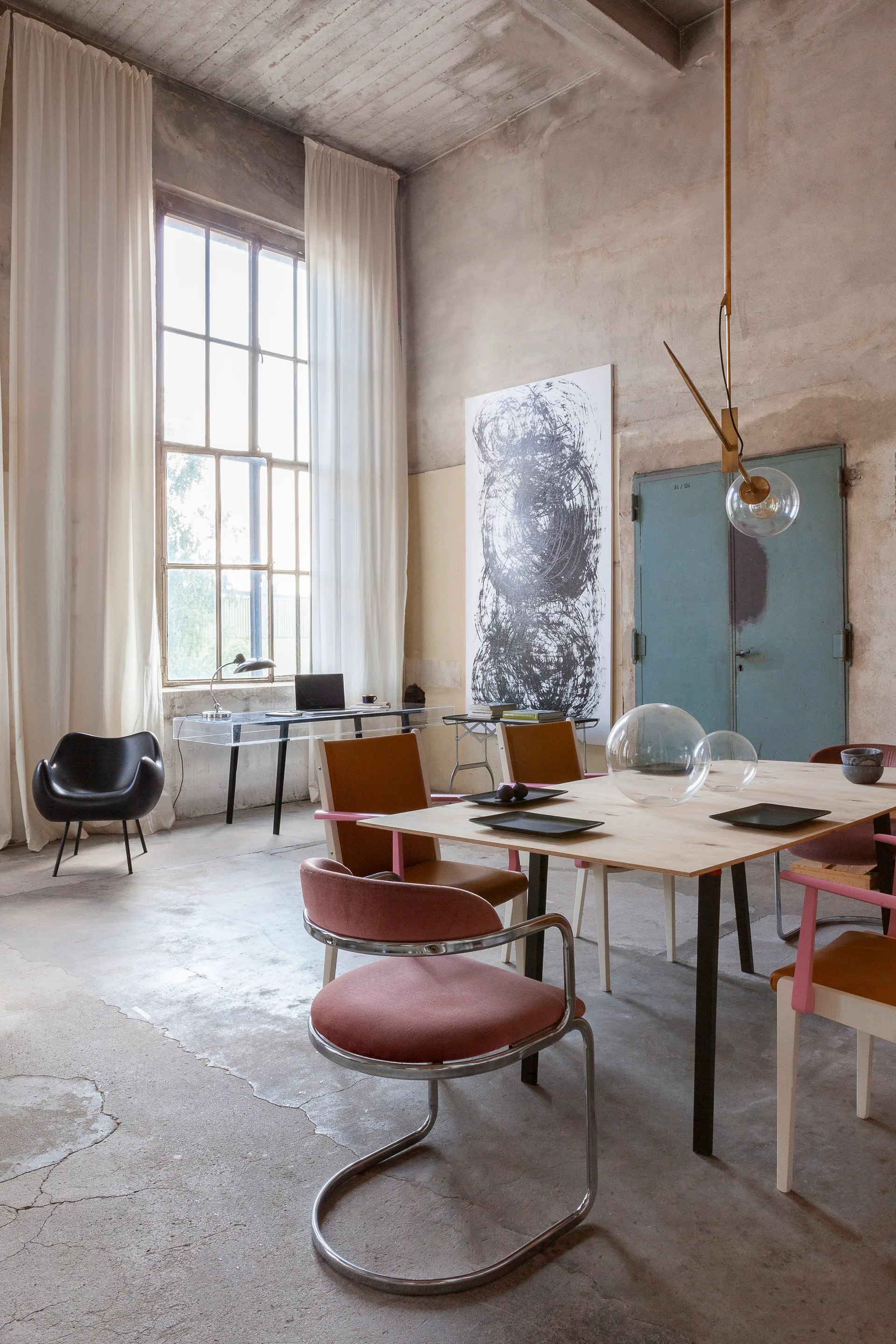 A spacious industrial loft features a light wood dining table with mixed chairs, an acrylic desk near a tall window, and exposed concrete walls.