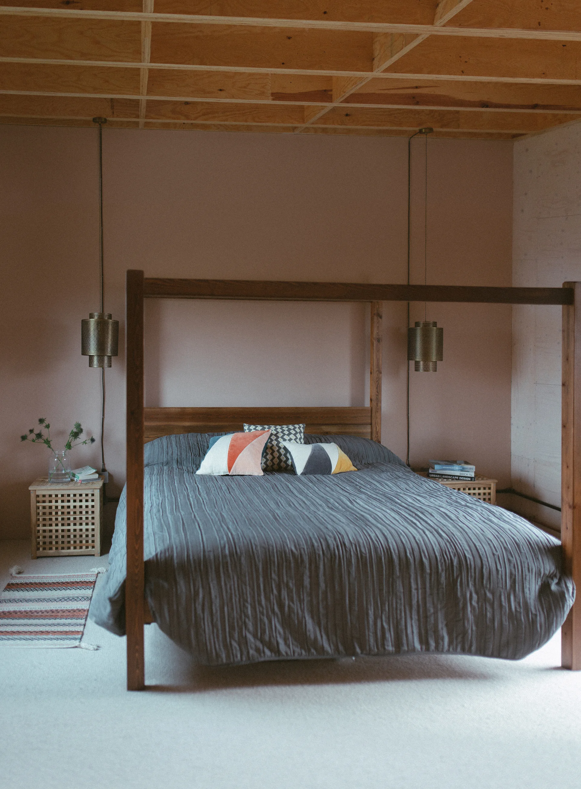 A wooden canopy bed with a grey textured duvet sits against a dusty pink wall under an exposed plywood grid ceiling in a bedroom.