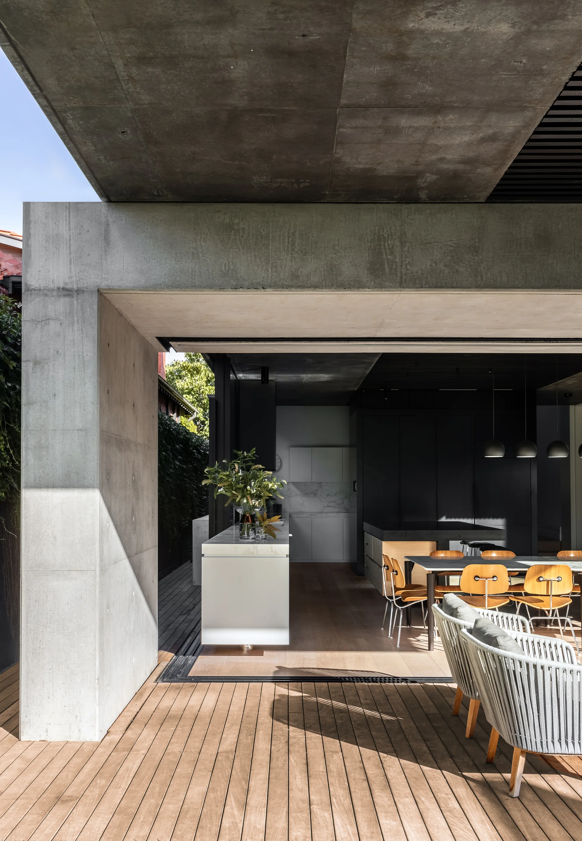 A modern indoor-outdoor kitchen features an exposed concrete structure, dark cabinetry, a white island, and a wooden deck.