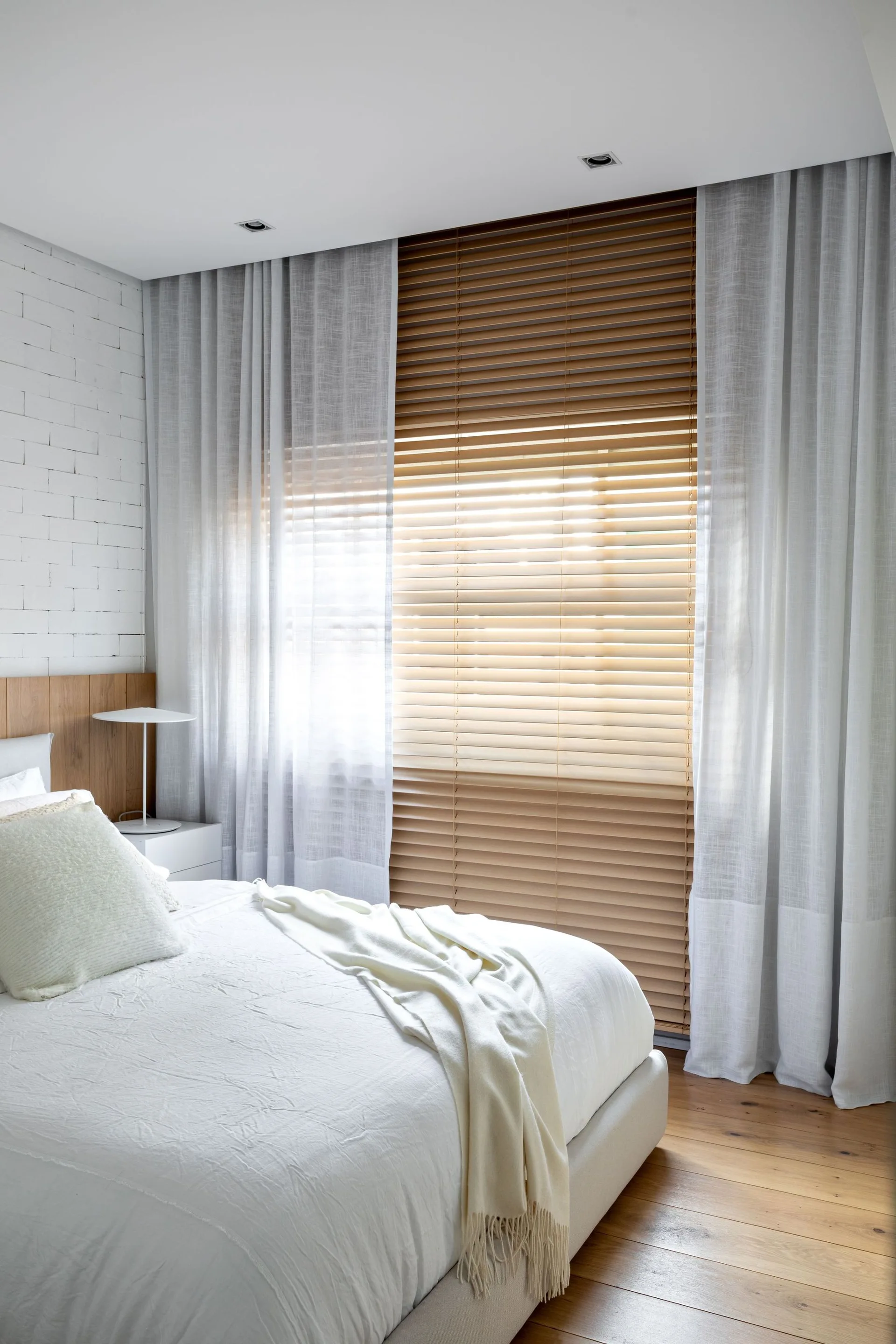 A minimalist bedroom featuring a white bed next to a painted brick wall, with warm wooden blinds layered under sheer white curtains.