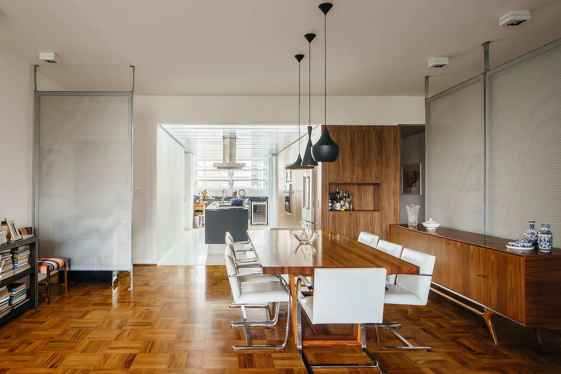 A modern dining room features a wooden table with white chairs, parquet flooring, black pendant lights, and a kitchen in the background.