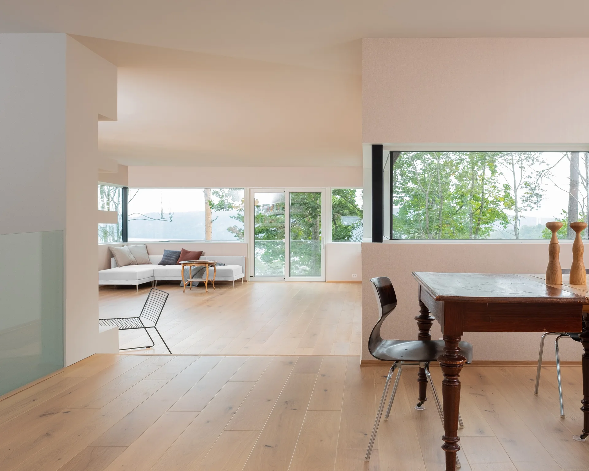 An open-concept living and dining area features light wood flooring, soft beige walls, a dark wood dining table, and large windows looking out to trees.