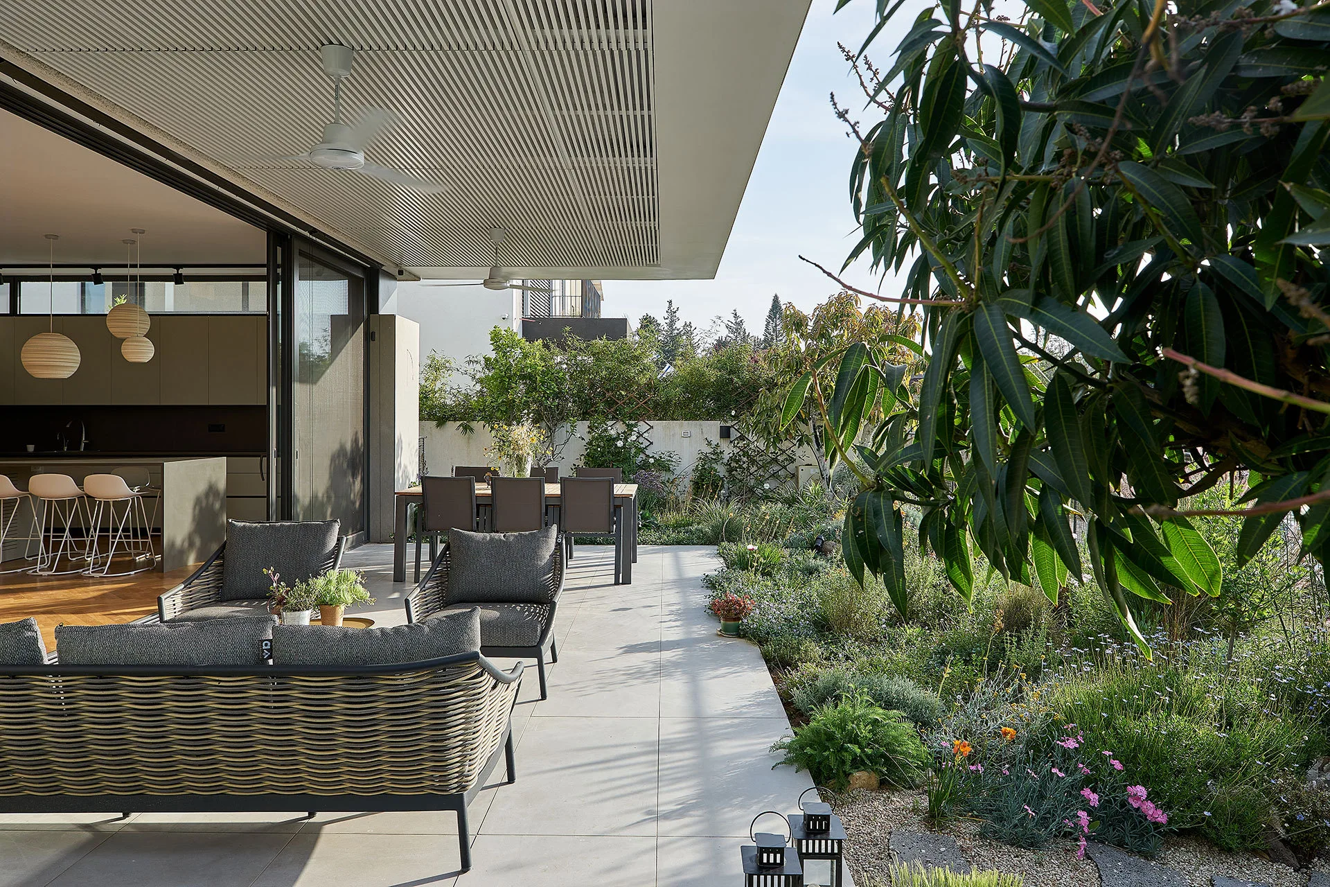 A contemporary tiled patio features gray woven seating and a dining table alongside a green garden, with glass doors opening to an indoor kitchen.