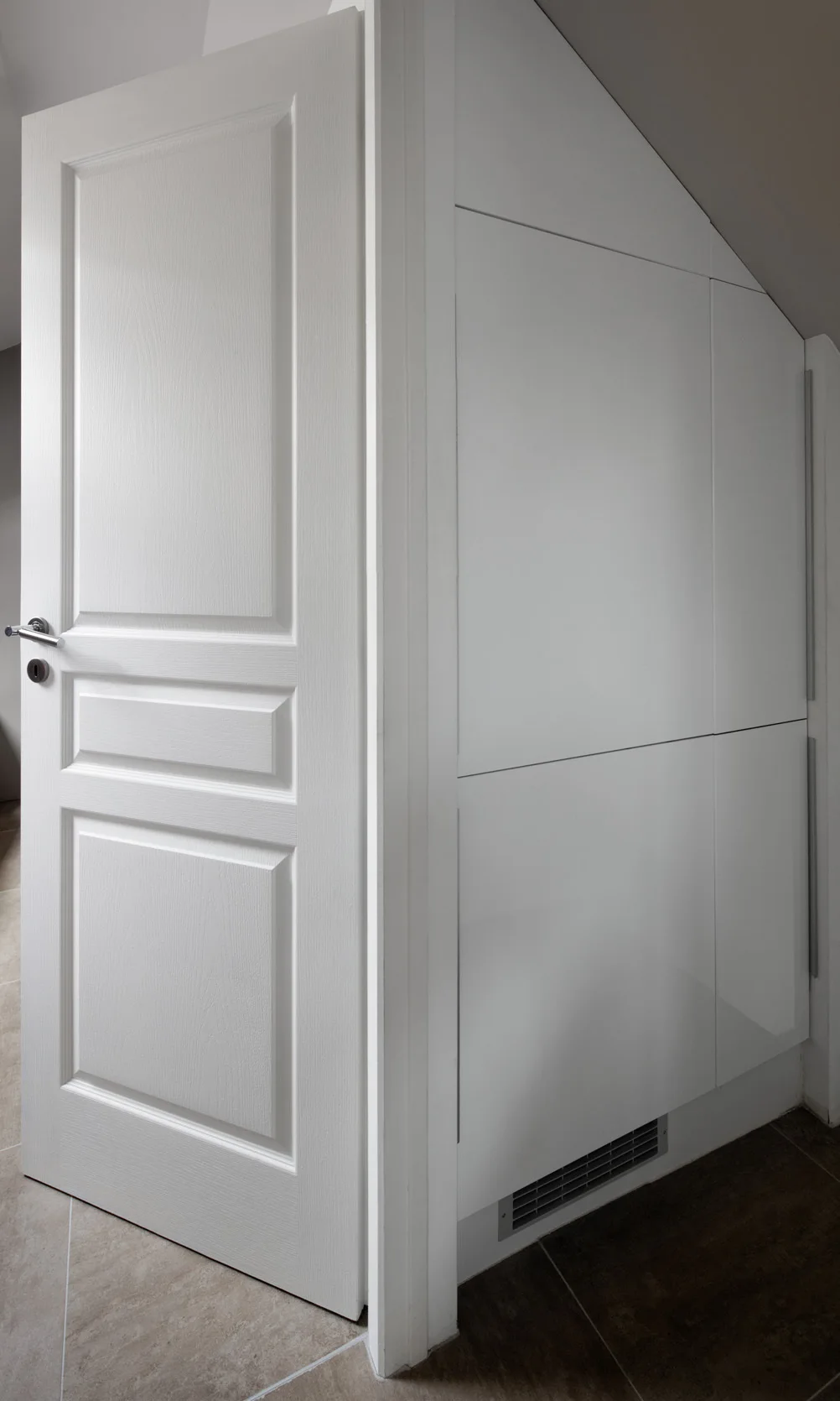 A white paneled door stands slightly open next to custom white minimalist cabinets fitted under a sloped ceiling.