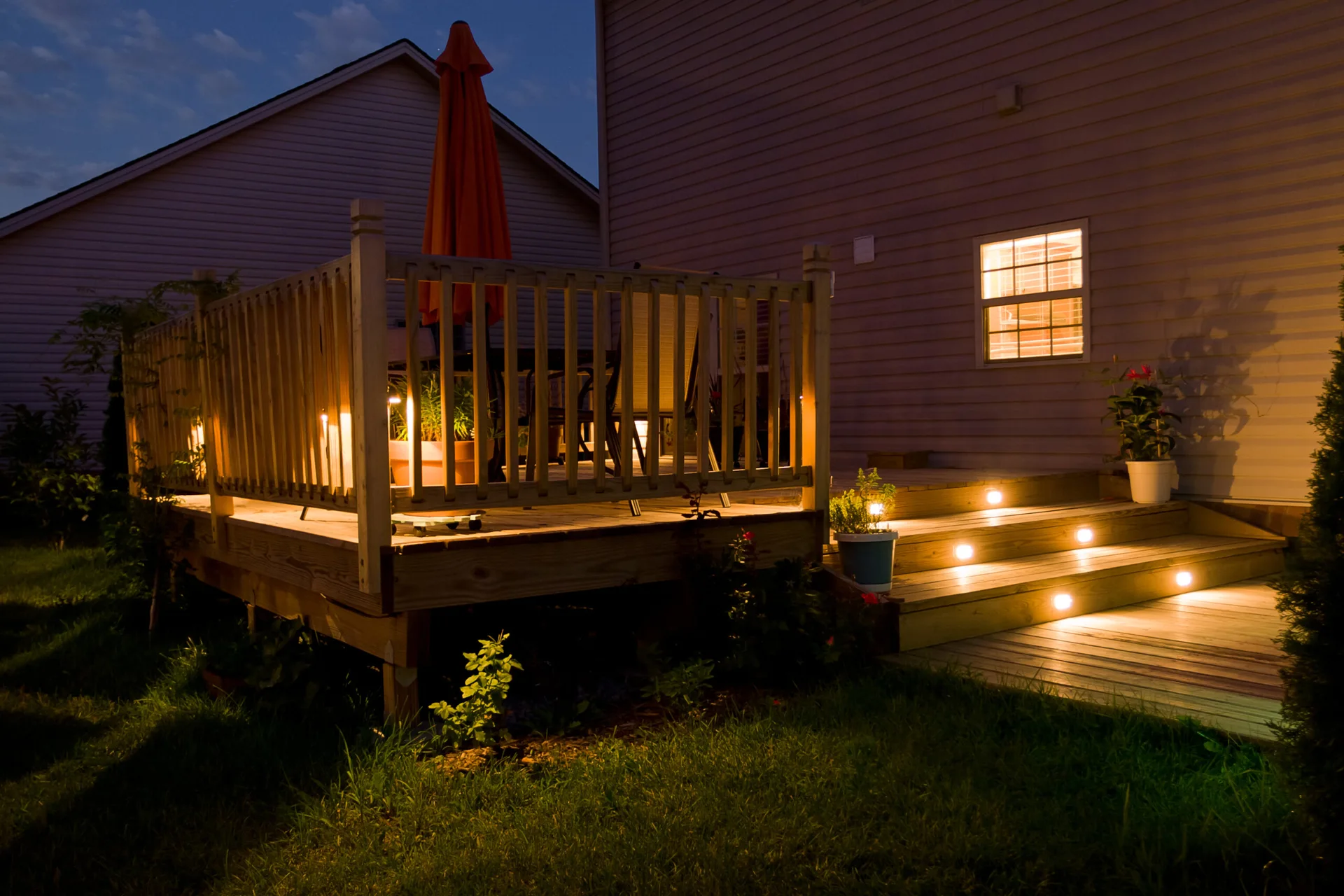 A wooden residential deck at night features illuminated wooden stairs with warm recessed lights, a railing, and a closed orange patio umbrella.