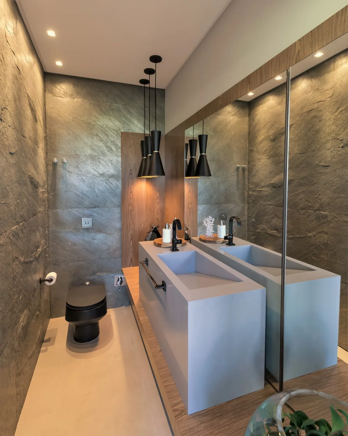 A modern powder room features textured stone walls, a light grey monolithic vanity sink, a black toilet, and three black cone pendant lights.