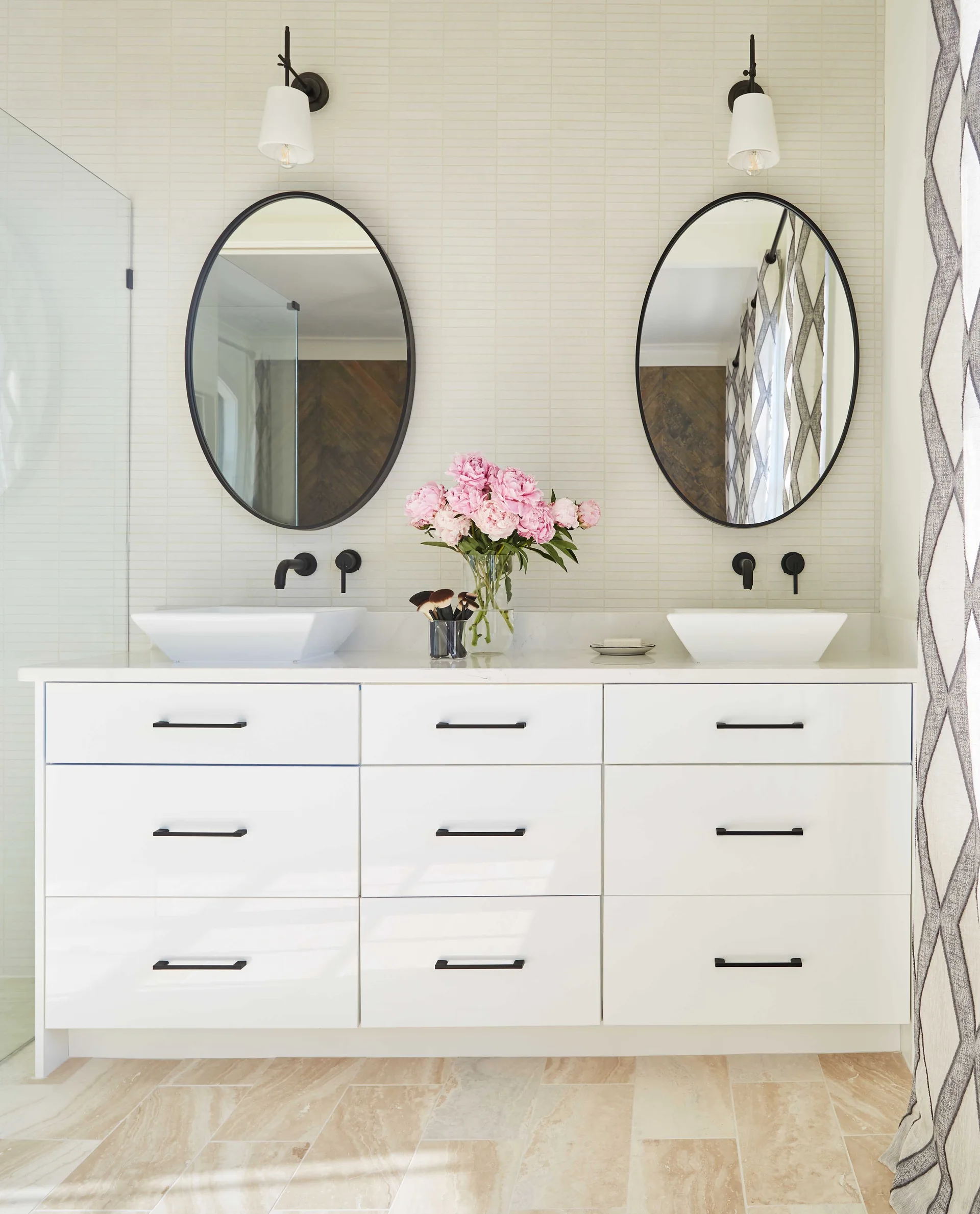 A bathroom features a white double vanity with vessel sinks, oval black-framed mirrors, wall-mounted faucets, and a light beige stacked tile backsplash.